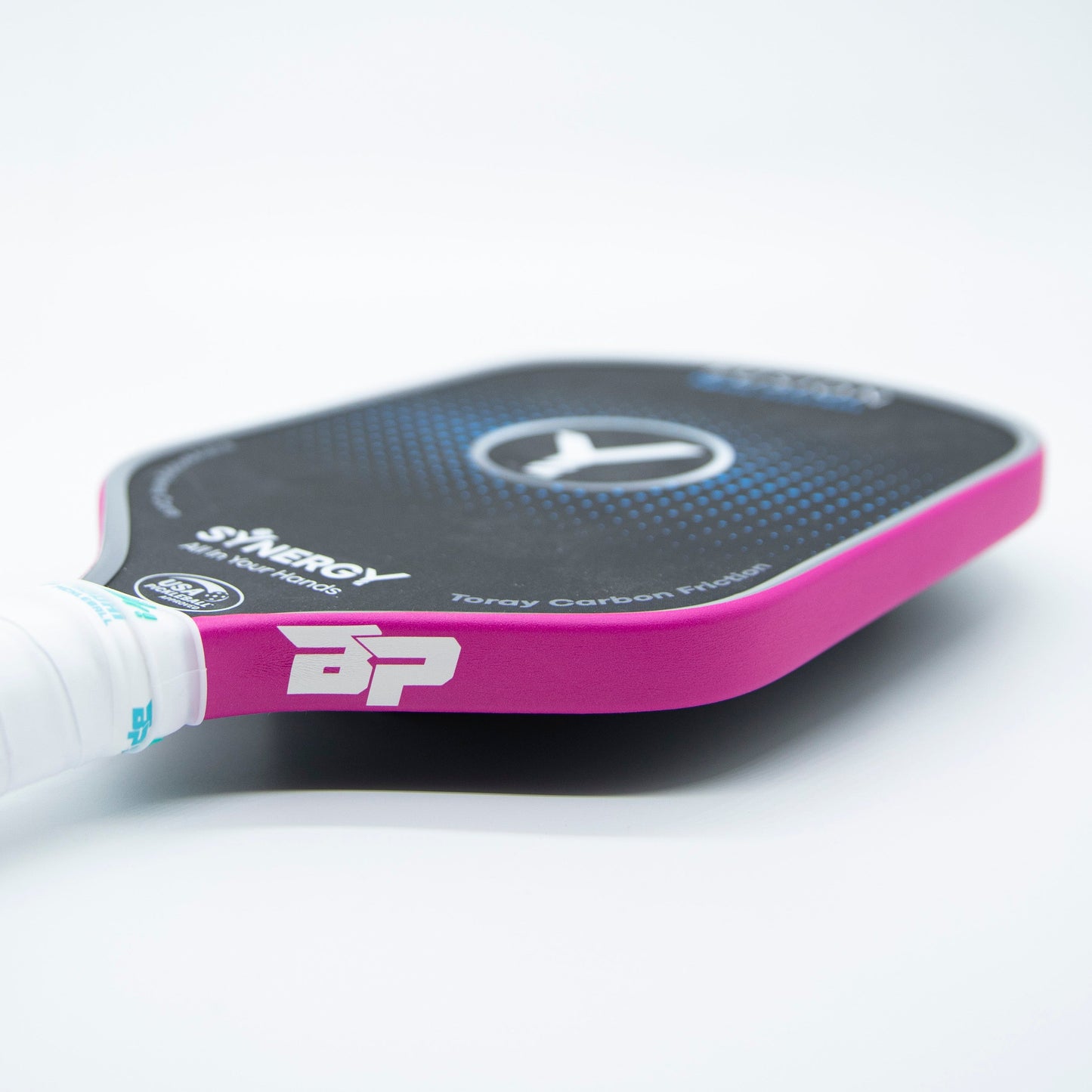 A close-up of a Bodhi Performance pickleball paddle with Premium Edge Tape - SOLIDS (black face, pink edge, white grip). BP and Synergy logos and “Tbray Carbon Paddle” text are visible; it rests on a plain white surface.