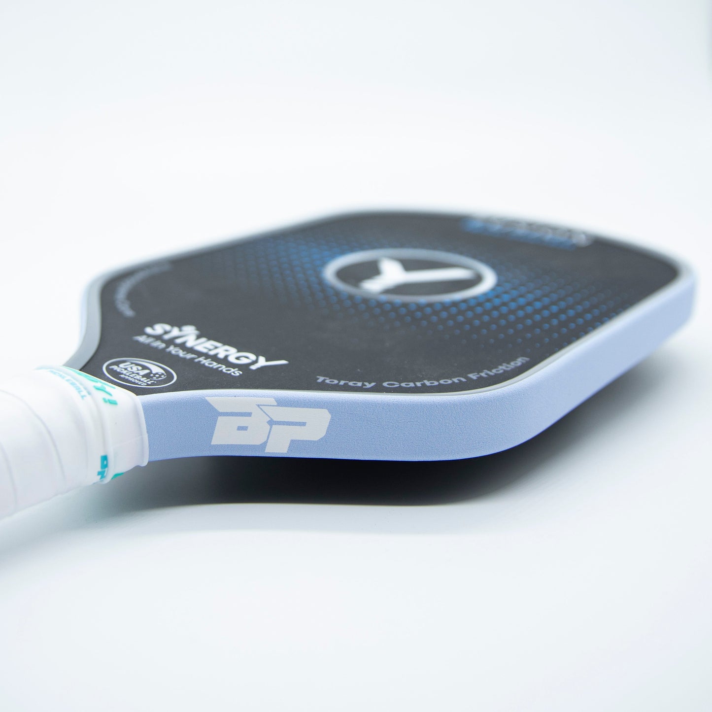 A close-up of a Bodhi Performance pickleball paddle with a white grip and Premium Edge Tape - SOLIDS for protection. The textured black and blue surface features the SYNERGY logo and “Toray Carbon Fiber” on a plain white background.