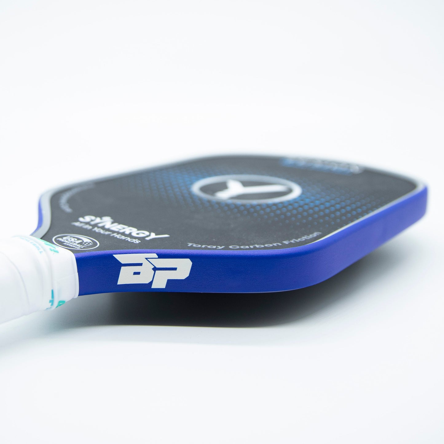 A close-up of a blue and black Bodhi Performance paddle with the “BP” logo, “Synergy,” and “Toray Carbon Fiber” on the face, a wrapped white grip, and Premium Edge Tape - SOLIDS for added protection, all on a white surface.