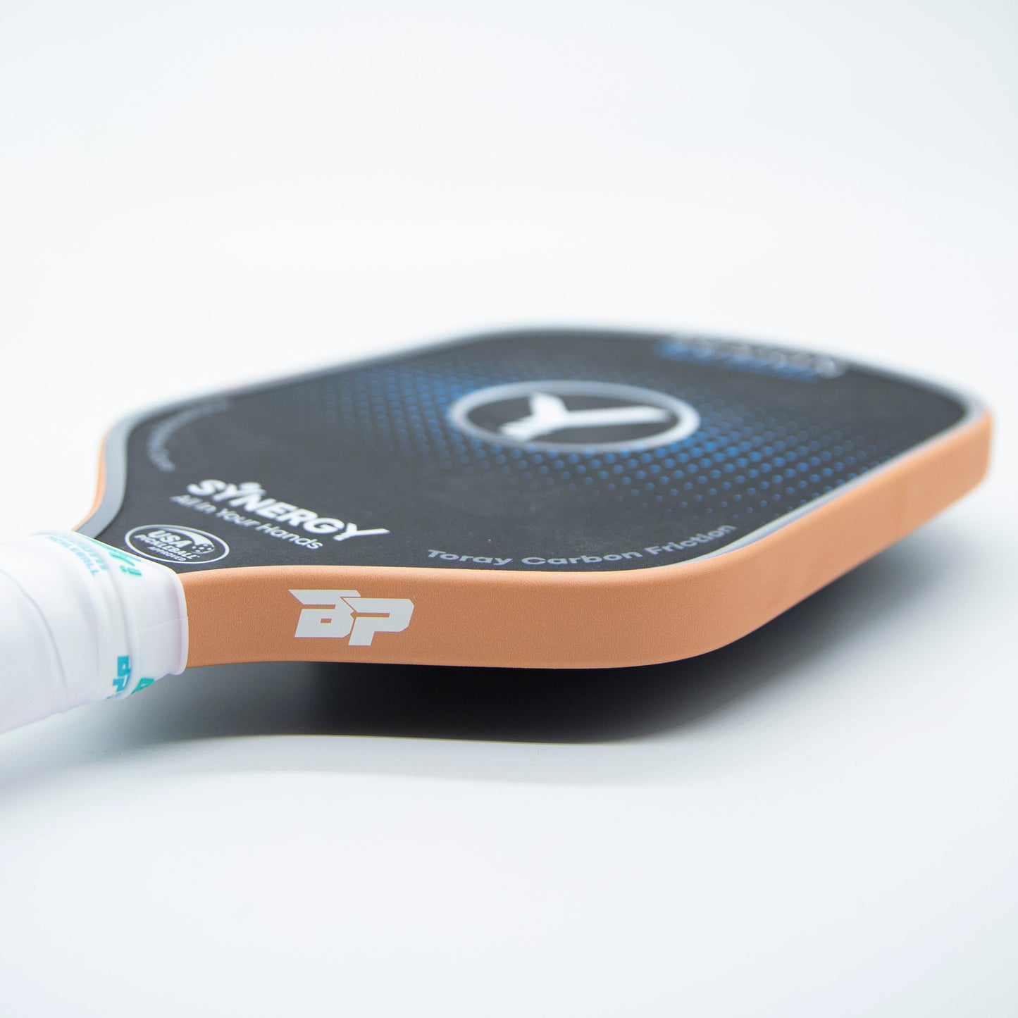 A close-up of a Bodhi Performance pickleball paddle shows a black face, orange edge, white grip, blue circular pattern, Synergy and BP branding, plus Premium Edge Tape - SOLIDS for added protection.