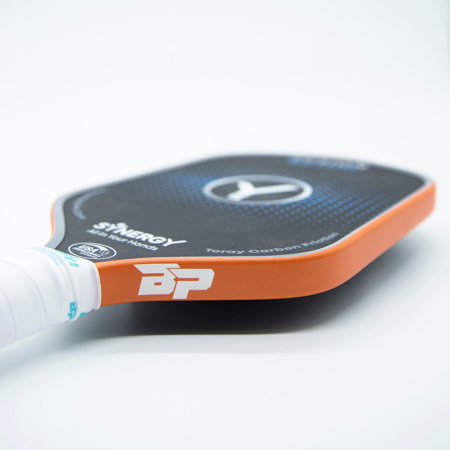 A close-up of a black and orange pickleball paddle with a white grip, featuring “SYNERGY,” a circular logo, and “Toray Carbon Fiber” along the edge, protected by Bodhi Performance Premium Edge Tape - SOLIDS for added durability.