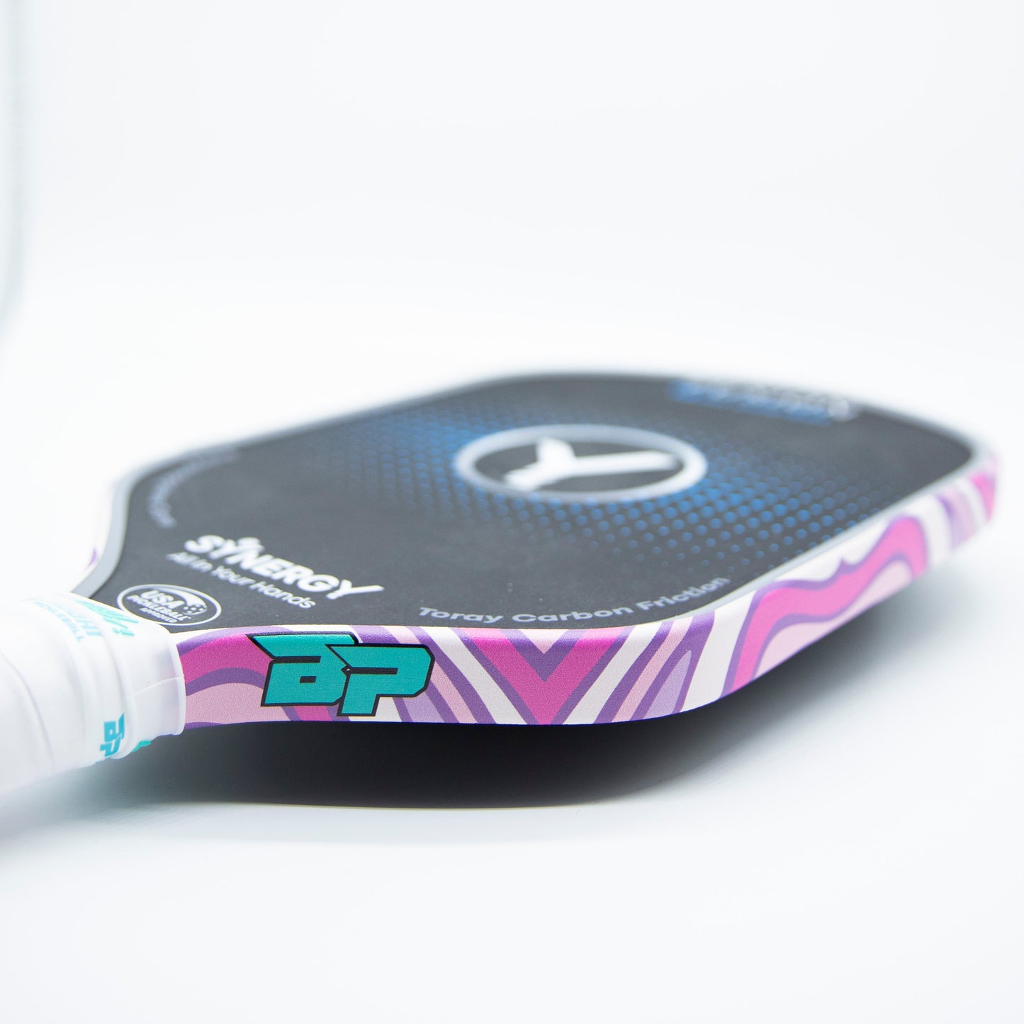 A close-up shows a Bodhi Performance paddle with a black face, white circular logo, purple and pink waves edged by Premium Edge Tape - PATTERNS, a wrapped white handle, and “BP” near the handle.