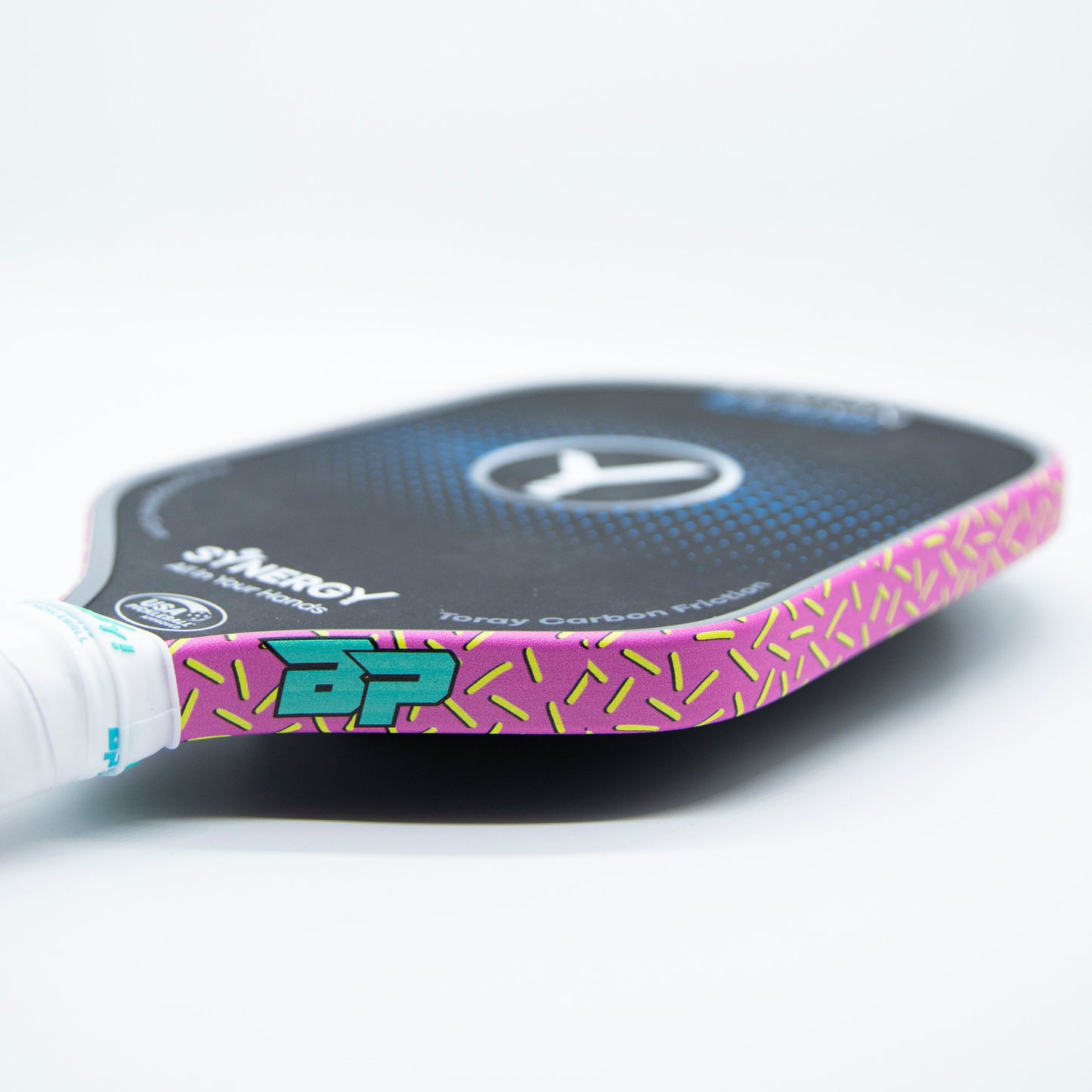 A close-up of a Bodhi Performance pickleball paddle highlights its Premium Edge Tape - PATTERNS in pink and yellow for added protection, a turquoise "BP" logo, white grip tape handle, branding, and circular design on a white surface.