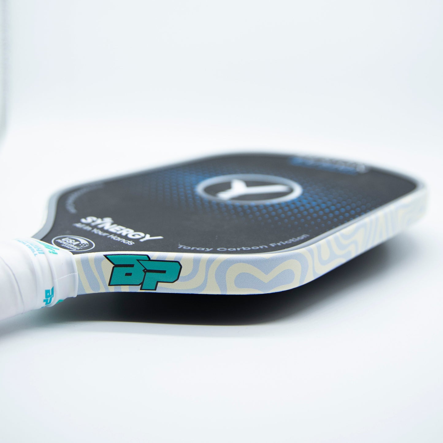 A close-up of a Bodhi Performance pickleball paddle with black textured face, blue dots, central logo, and Premium Edge Tape – PATTERNS for protection. The handle has white grip tape with a turquoise BP logo.