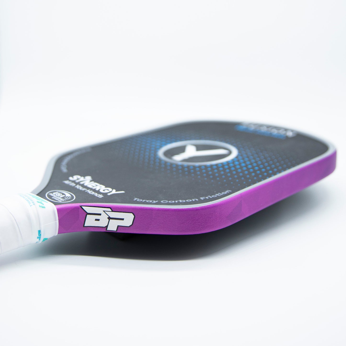A close-up of the Bodhi Performance Premium Edge Tape - PATTERNS shows purple edge guard protection, a white grip, and a black face with blue and white circles. Branding and product info are visible near the handle on this must-have pickleball accessory.