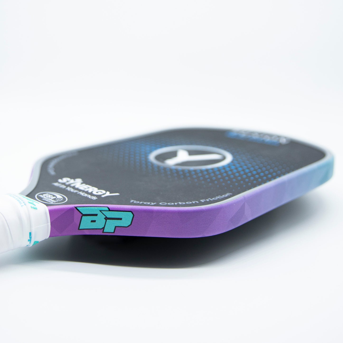 A close-up of a pickleball paddle featuring Bodhi Performance Premium Edge Tape - PATTERNS adds extra protection, with a black face, light blue and purple gradient edges, white grip, geometric design, branding, and model details.