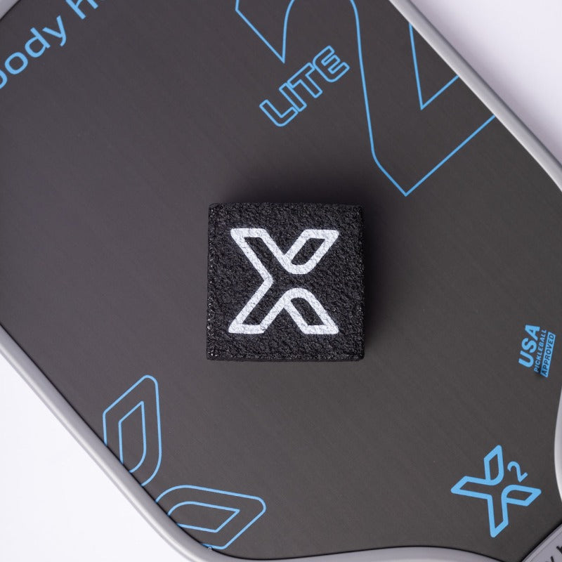 A close-up of the body helix Pickleball Paddle Eraser and Refresher, a black rectangular eraser with a white “X” logo, resting on a dark gray fitness board featuring blue outlines, “LITE,” “USA,” and extra “X” logos for paddle maintenance.