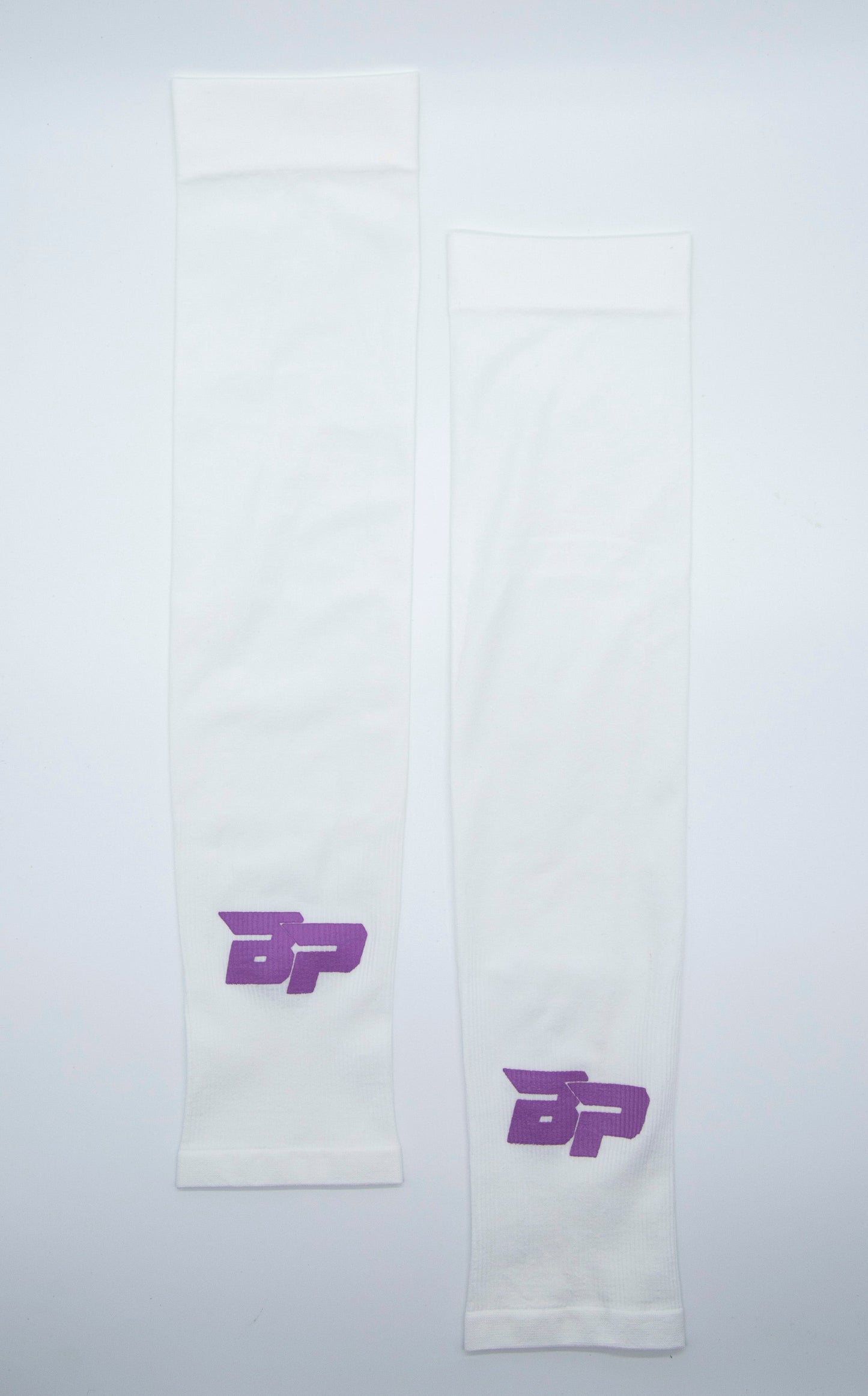 Two white Bodhi Performance arm sleeves are laid flat on a white surface. Each Performance Arm Sleeve displays a purple stylized BP logo near one end, with even lighting highlighting their smooth, moisture-wicking texture.
