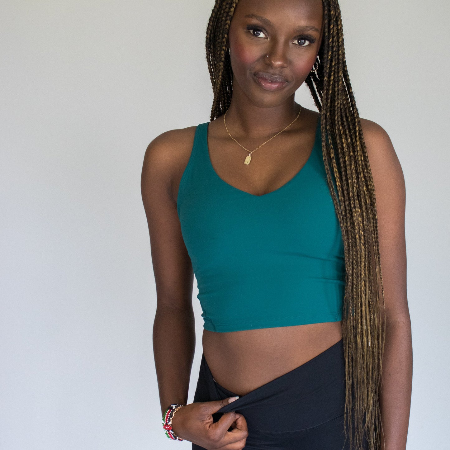 A woman with long, braided hair stands against a plain background in a teal DYM Athletics Flex V-Neck Bra with Scoop Back, smiling gently as she slightly pulls down her black bottoms, exuding relaxed confidence and comfort.