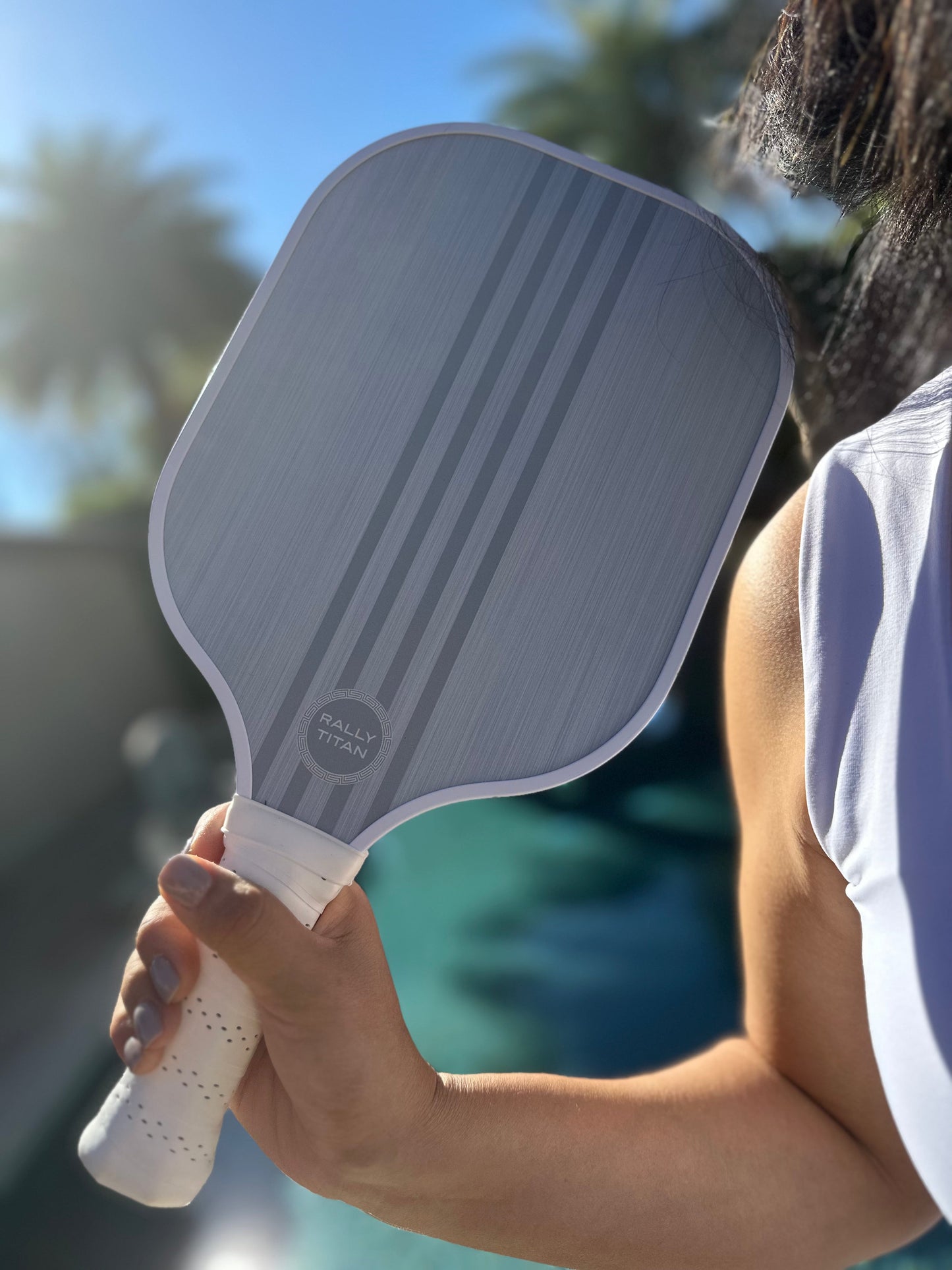A person in a white sleeveless top holds the Rally Titan Pickleball Grizo | 10MM paddle with a T300 textured carbon fiber surface and white grip near a sunlit pool, with palm trees and a clear sky blurred in the background.