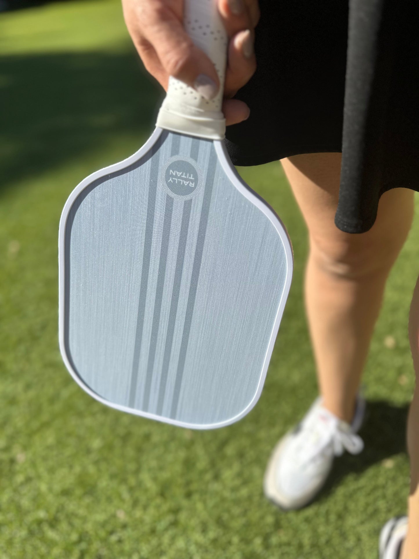 A person holds a Rally Titan Pickleball Grizo 10MM paddle with a T300 textured carbon fiber surface, standing on green grass. Only their hand, lower legs, black skirt, and white sneaker are visible in the sunlight.