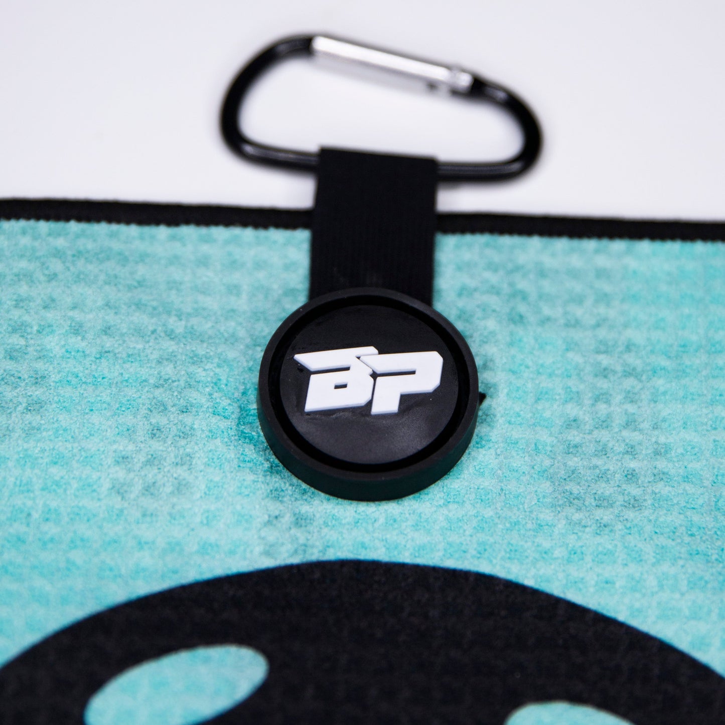 A close-up of a teal Bodhi Performance Pickleball Towel shows its textured fabric and a black circular tag with raised white "BP" letters and a wing, attached by a strap with a metal carabiner clip.