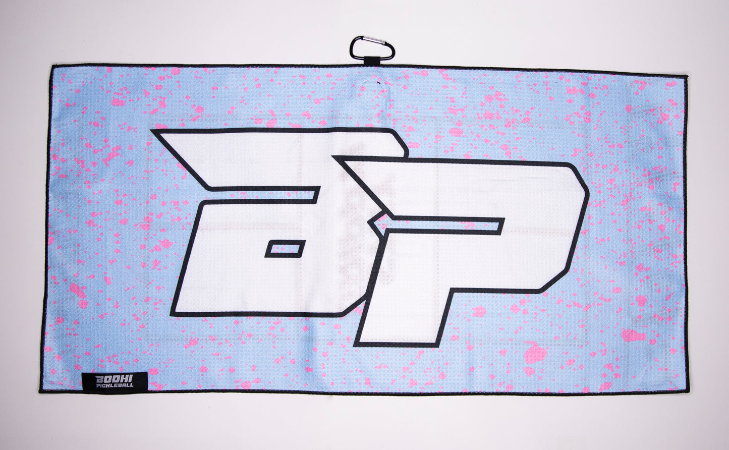 The Bodhi Performance Pickleball Towel is a light blue rectangular microfiber towel with large white “ap” letters outlined in black, pink paint splatters, a sturdy black metal clip at the top, and a small black label at the bottom left.