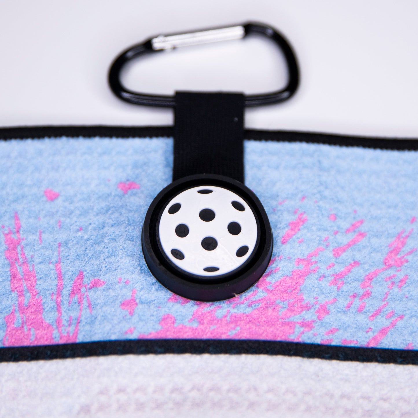 Close-up of the Bodhi Performance Performance Pickleball Towel in light blue and pink with a black-bordered edge, black strap, white six-hole disc, and carabiner—an essential accessory for any pickleball player.