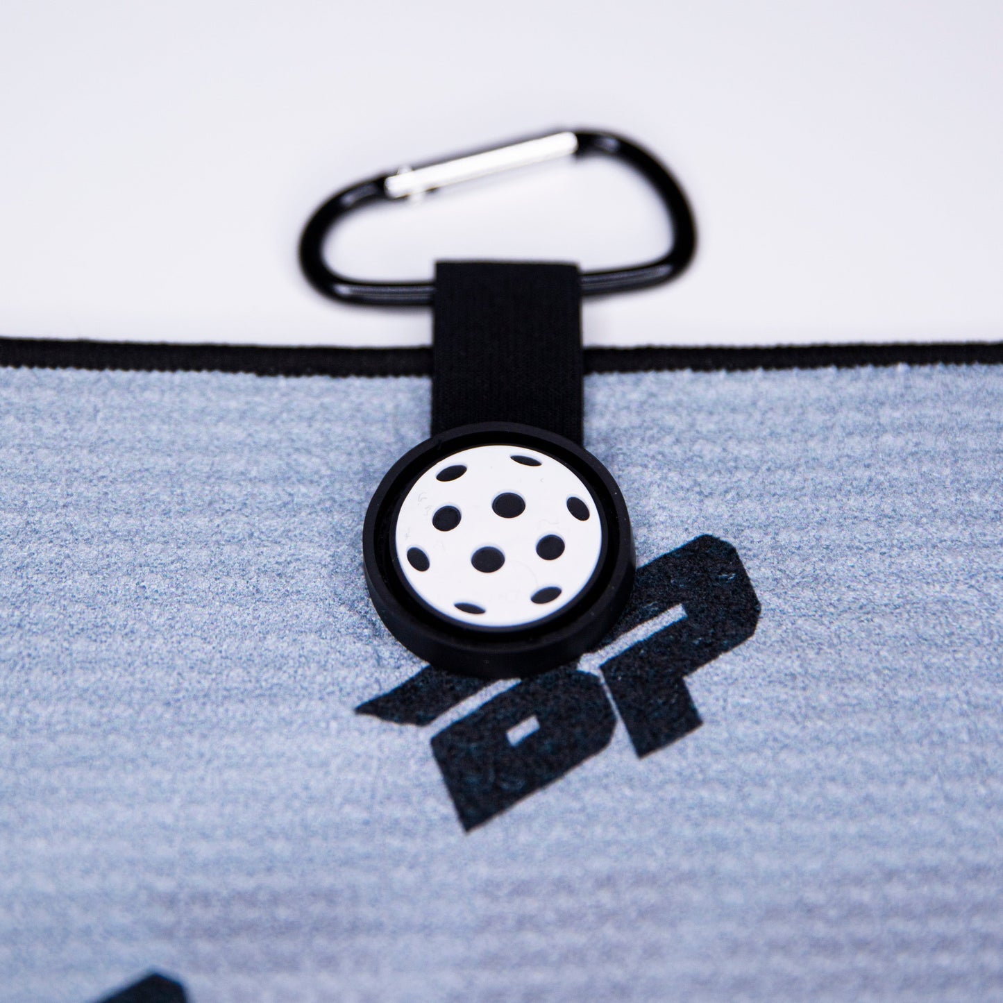 A close-up of the Bodhi Performance Performance Pickleball Towel shows its gray textured microfiber fabric with a black and white perforated circular clip, a carabiner, bold black logo details, and a black elastic loop—perfect for pickleball accessories.