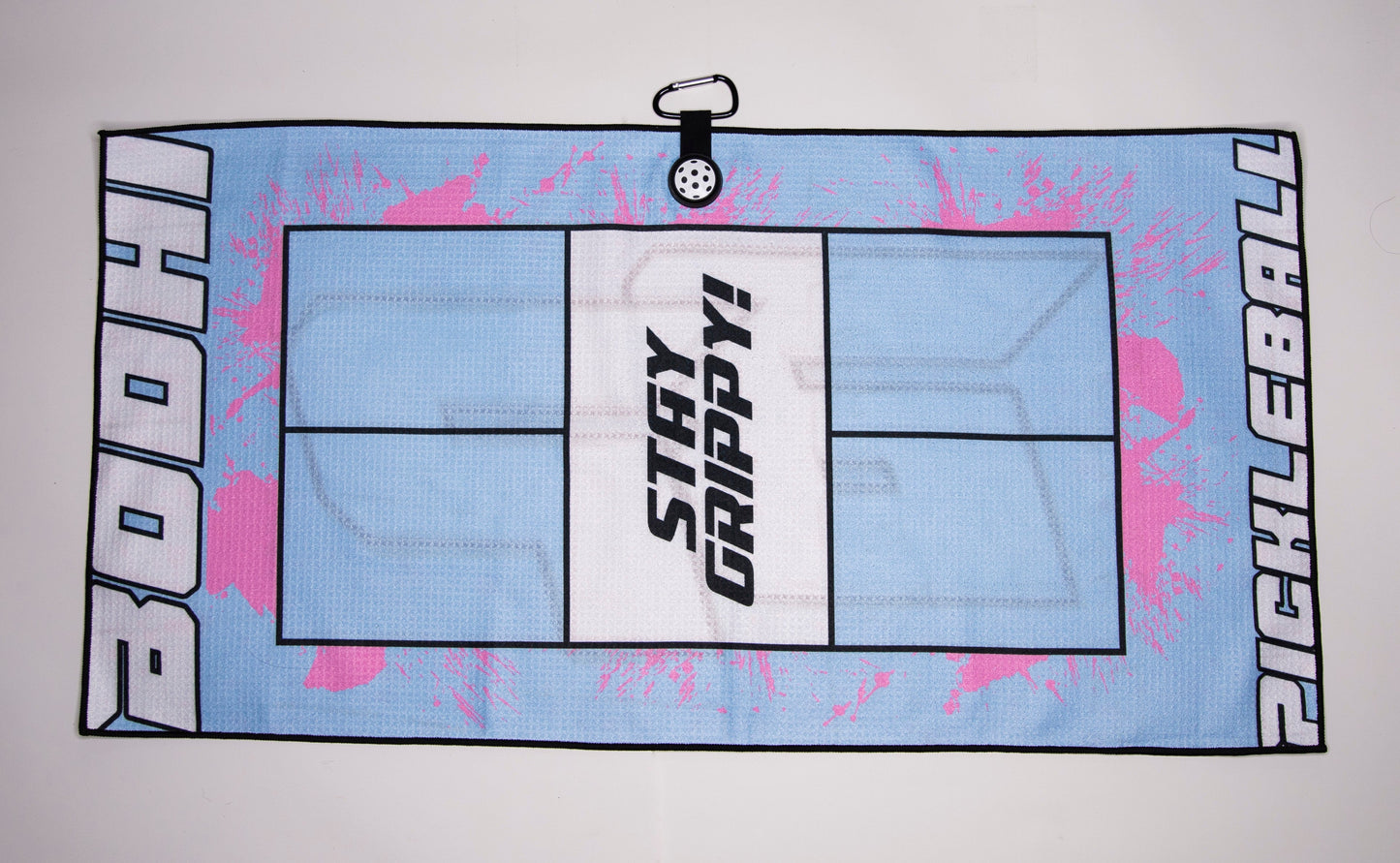 The Bodhi Performance Pickleball Towel is light blue microfiber with pink splatters, a court outline, and “STAY GRIPPY!” in bold. “BODHI” and “PICKLEBALL” are on each side. Includes a black clip topped with a white perforated pickleball.
