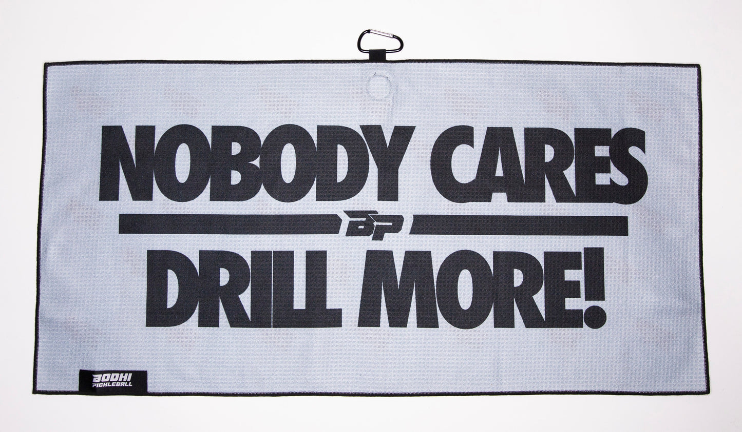 The Bodhi Performance Pickleball Towel is a rectangular gray microfiber towel with a black border, bold “NOBODY CARES DRILL MORE!” text, carabiner clip, small logo, and label—a must-have accessory for pickleball players.