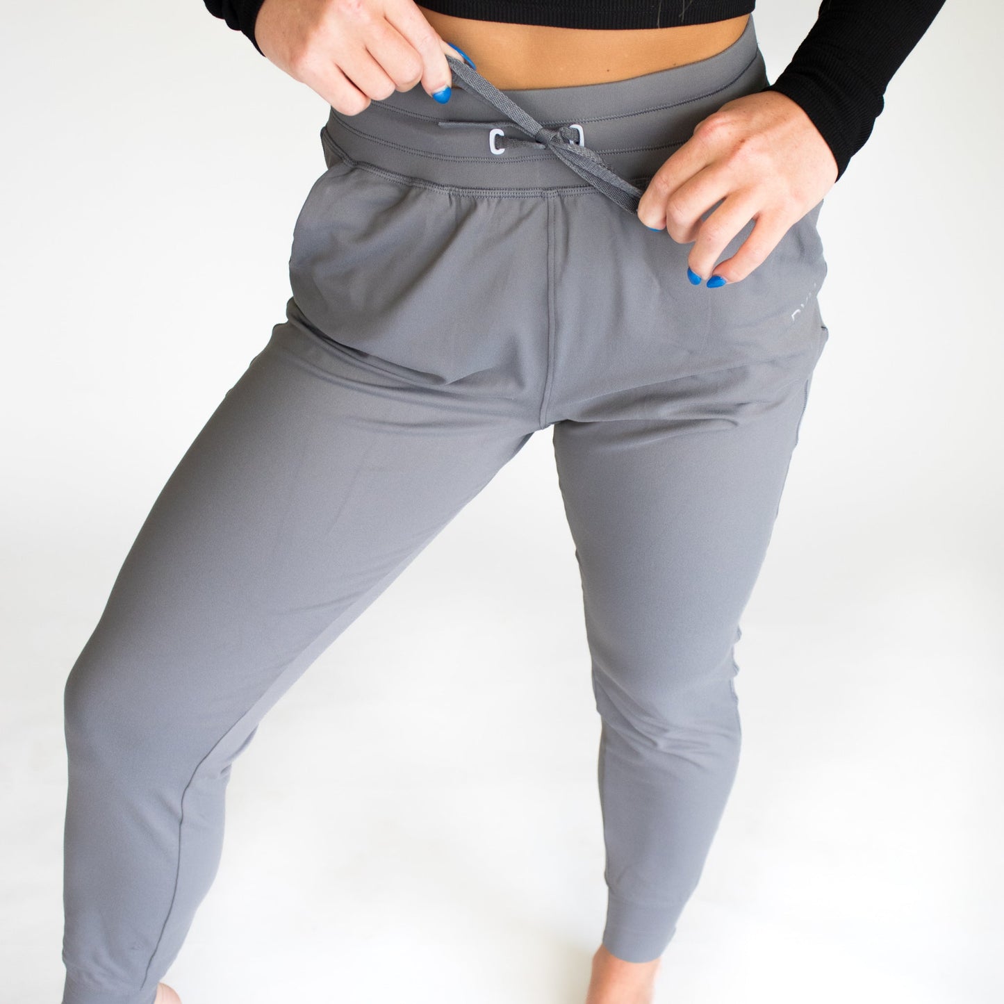 A person with blue-painted nails adjusts the waistband of DYM Athletics' Performance Pro Lightweight Active Jogger in light gray. Barefoot and in a long-sleeve black top, they stand against a white backdrop, highlighting this stylish athletic wear.