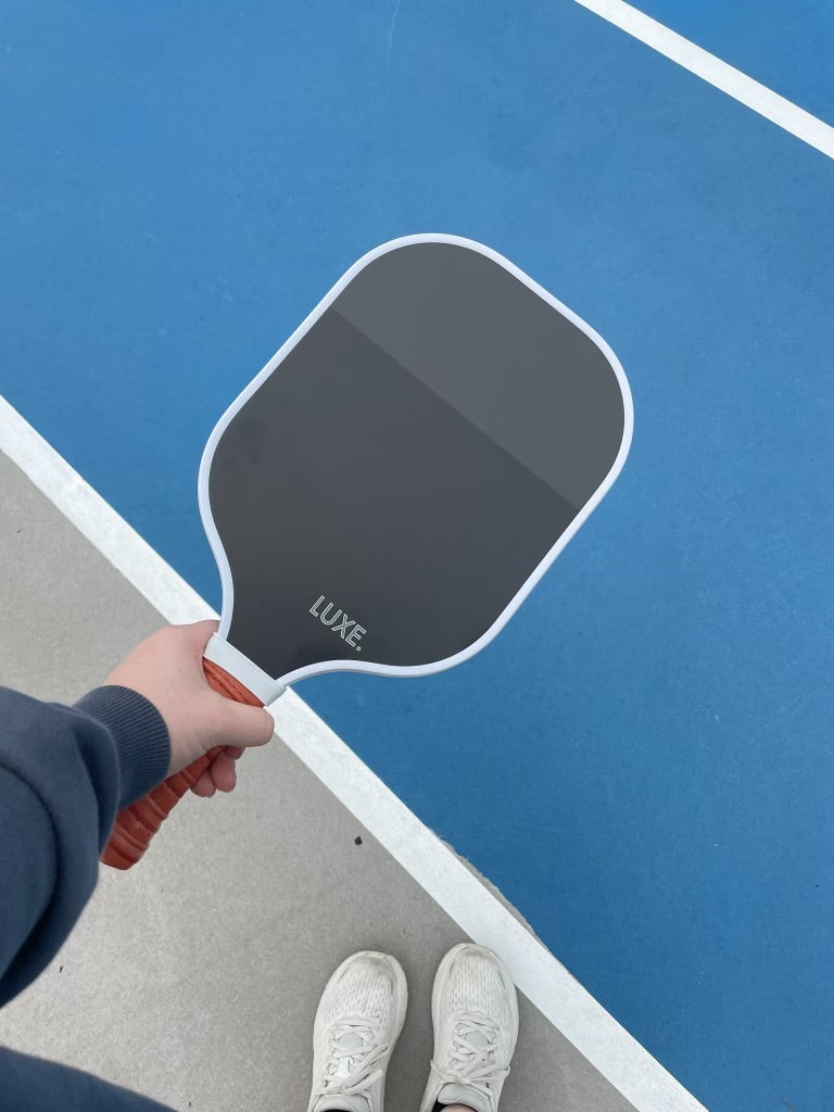 A person stands on a blue and gray court holding the LUXE Pickleball Dipped - Leisure paddle, which features a black face, brown handle, and honeycomb polymer core. They wear white sneakers and a dark sleeve on their arm.