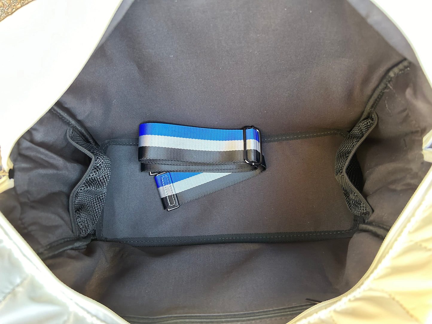 A close-up of the inside of Runway Athletics' The Quilted Puffer Pickleball Tote - White shows black fabric lining, two mesh side pockets, and a blue, gray, and black striped adjustable shoulder strap with a black plastic clip at the bottom.