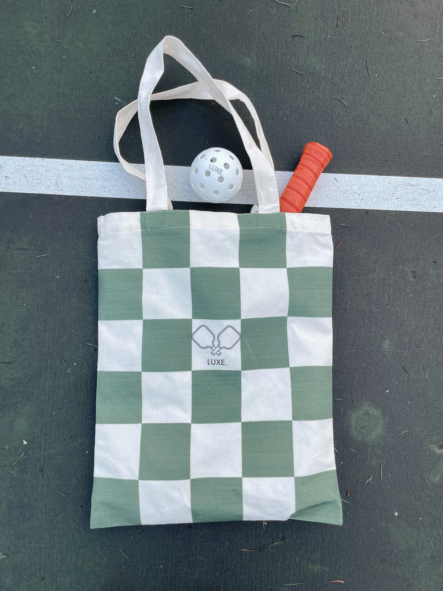 A green and white checkered Tote Bag from LUXE Pickleball, featuring two overlapping tennis rackets on the front, rests on a dark court. A white pickleball and an orange-handled paddle are visible from the bag’s opening.