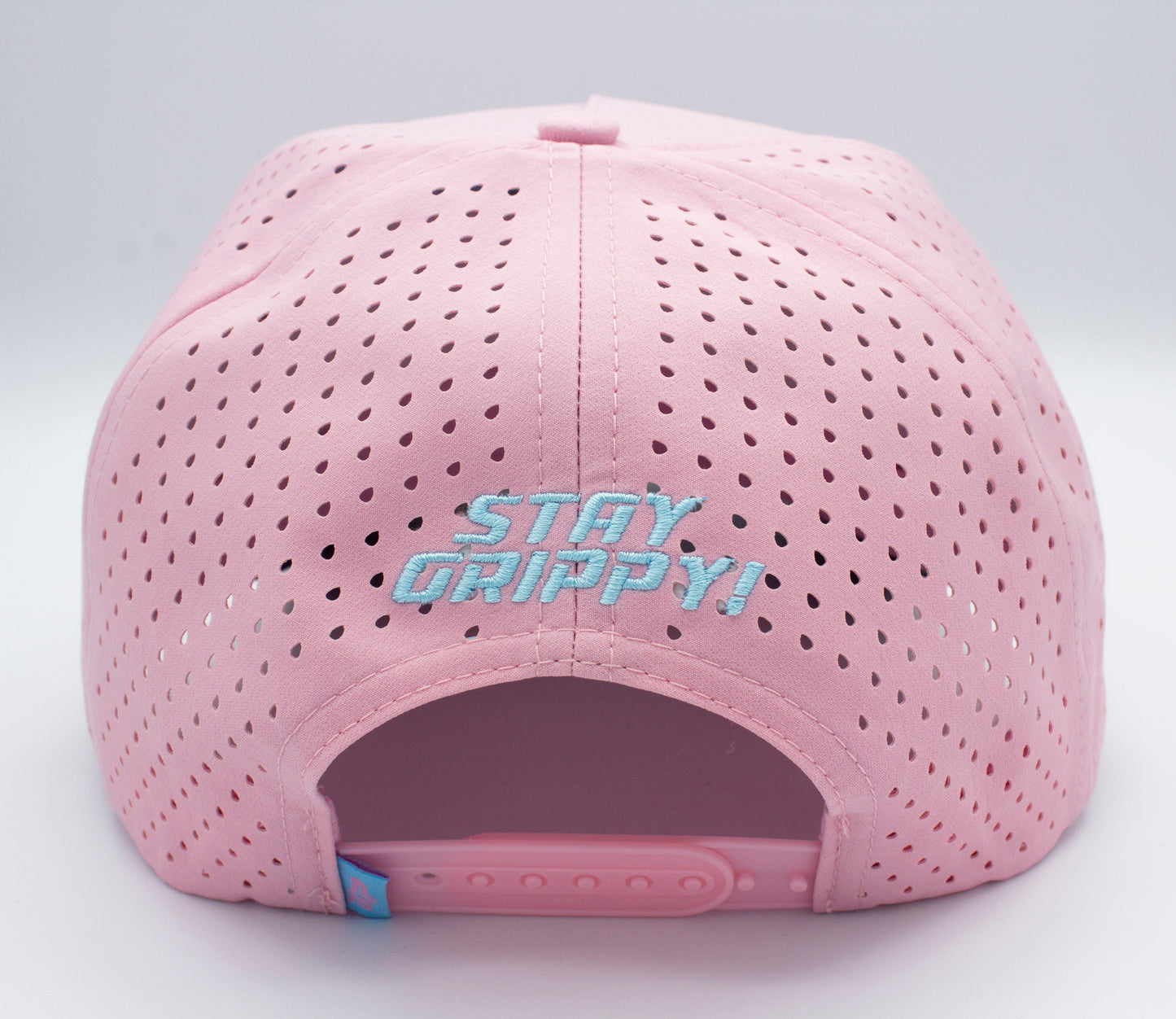 The ICON Snapback by Bodhi Performance is a light pink pickleball hat shown from the back, with small ventilation holes and a matching adjustable strap. Above the opening, “STAY GRIPPY!” is embroidered in white and blue on pink fabric. Plain white background.