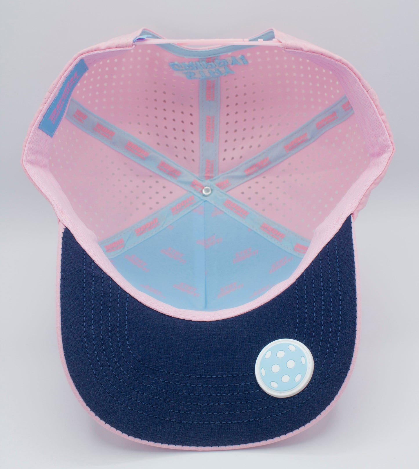 A photo shows the inside of the Bodhi Performance ICON Snapback, featuring a light pink interior with small perforations, blue inner panels, a navy blue brim, and a round pin with a light blue polka dot design on the left side.