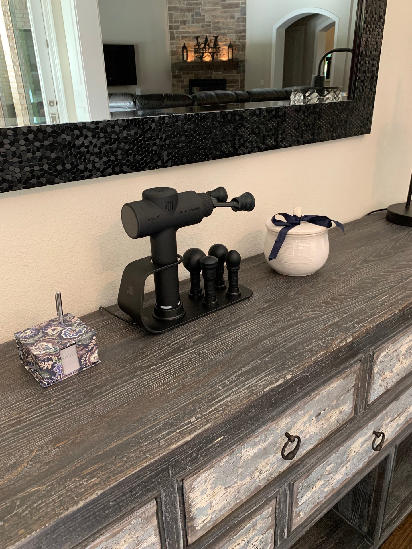 A dark wooden dresser holds an iReliev MVP+ PowerUp Charging Station, a tissue box, a white ceramic container with a blue ribbon, and a large black framed mirror reflecting a living room with a brick fireplace.