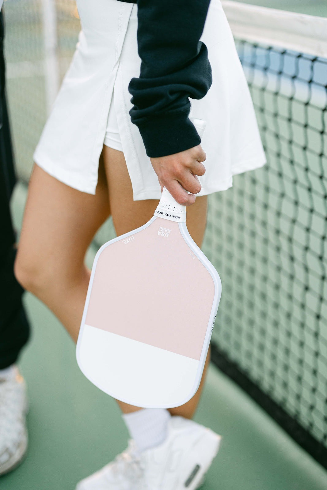 A person in a white skirt and black long-sleeve top holds the LUXE Pickleball Dinker, a USA Pickleball Approved light pink and white carbon fiber paddle, by their side on an outdoor court with the net visible. Only their lower body and hand are shown.
