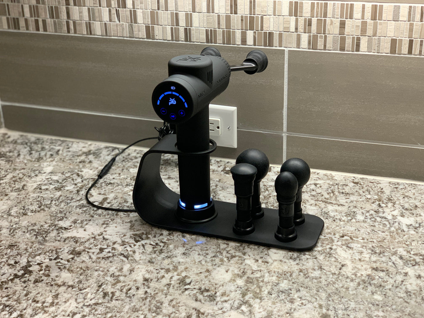 A black iReliev MVP-2000 Percussion Massager with digital display and blue LED lights sits docked in the MVP+ PowerUp Charging Station, which holds three massage heads. Tiled walls and an electrical outlet are visible in the background.