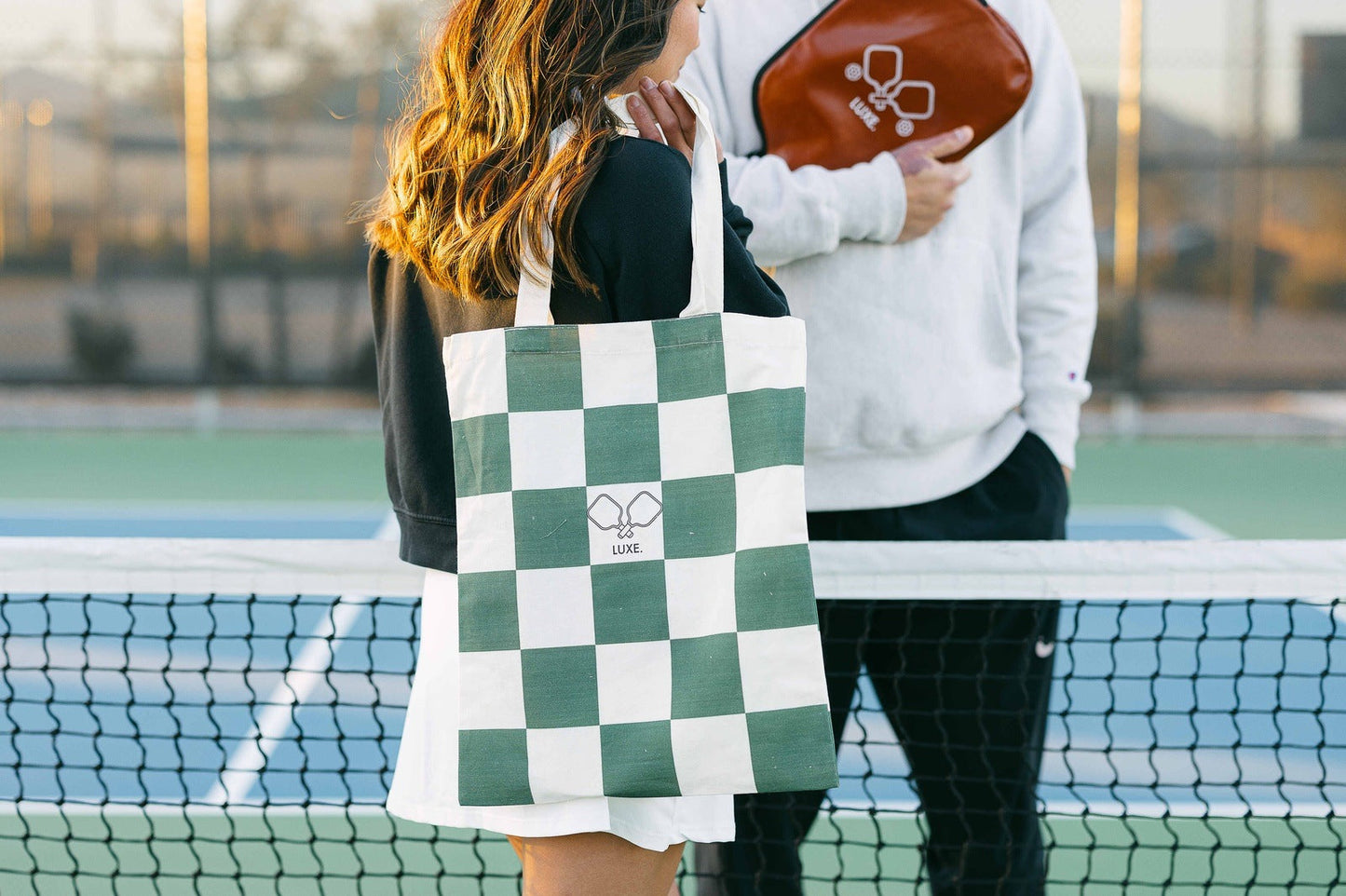 A woman stands on a tennis court holding the green and white checkered LUXE Pickleball Tote Bag with a small racket and ball illustration, while next to her, someone in a white sweatshirt holds a brown pouch with a white tennis racket design.
