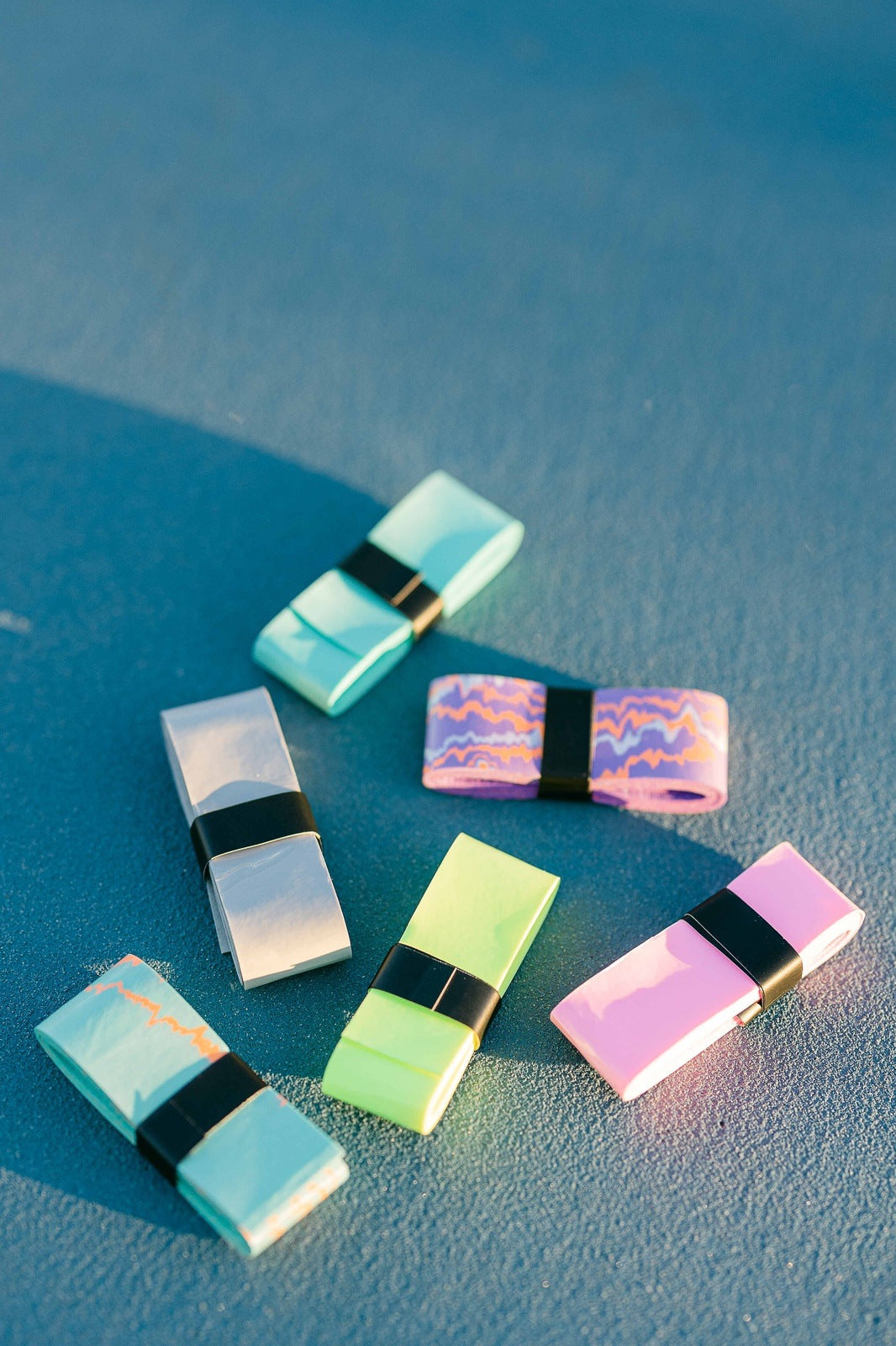 Six LUXE Pickleball Paddle Grips in pastel blue, green, pink, and silver—some with wavy multicolored patterns and black center bands—are arranged on a blue surface under sunlight, casting soft shadows.