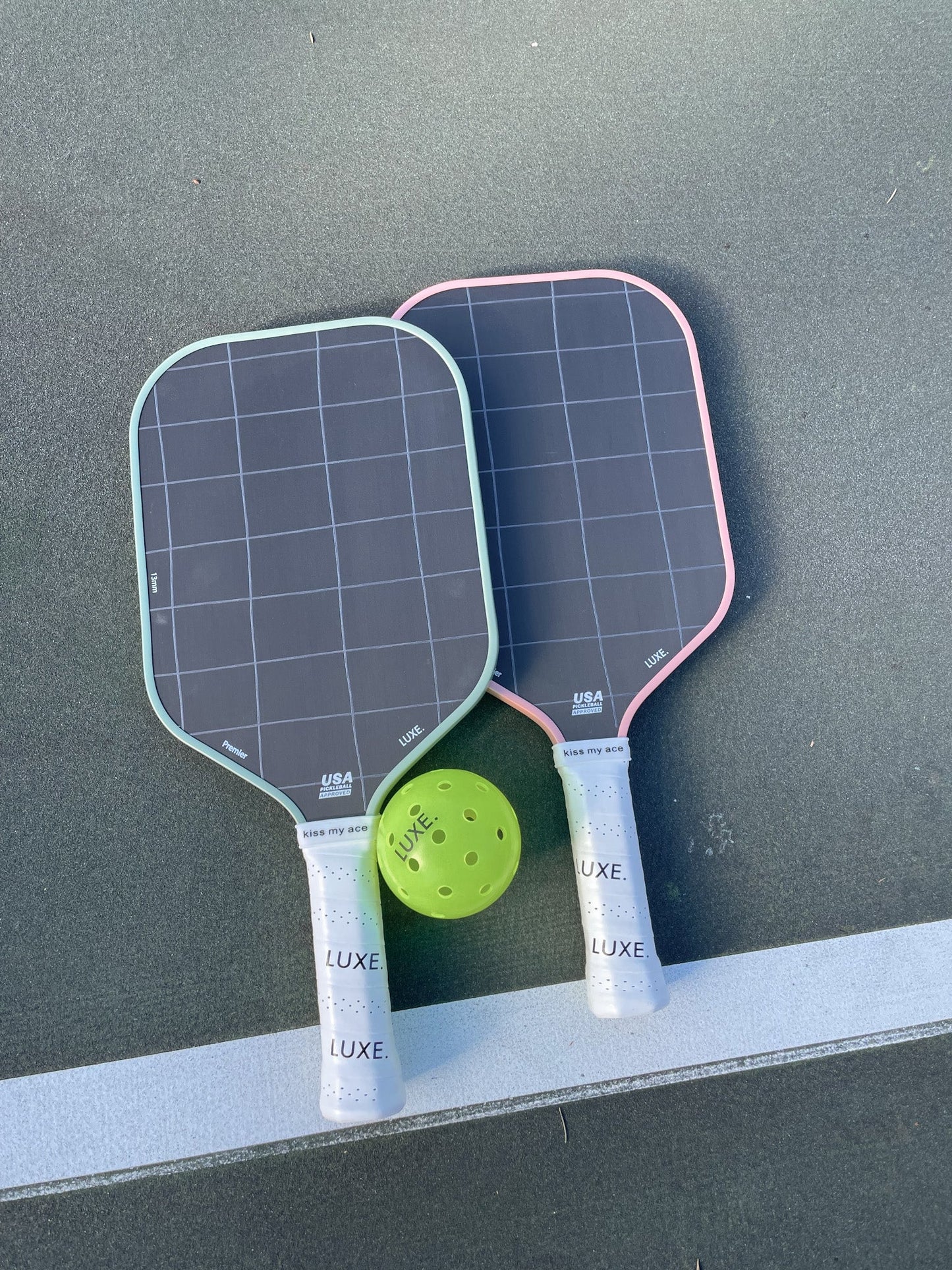 Two black LUXE Pickleball Premier paddles made with Toray t700 carbon fiber feature white grid patterns and “LUXE.” grips. Resting on a green court with a neon green ball, the paddles show pastel green and pink edges at the handles.