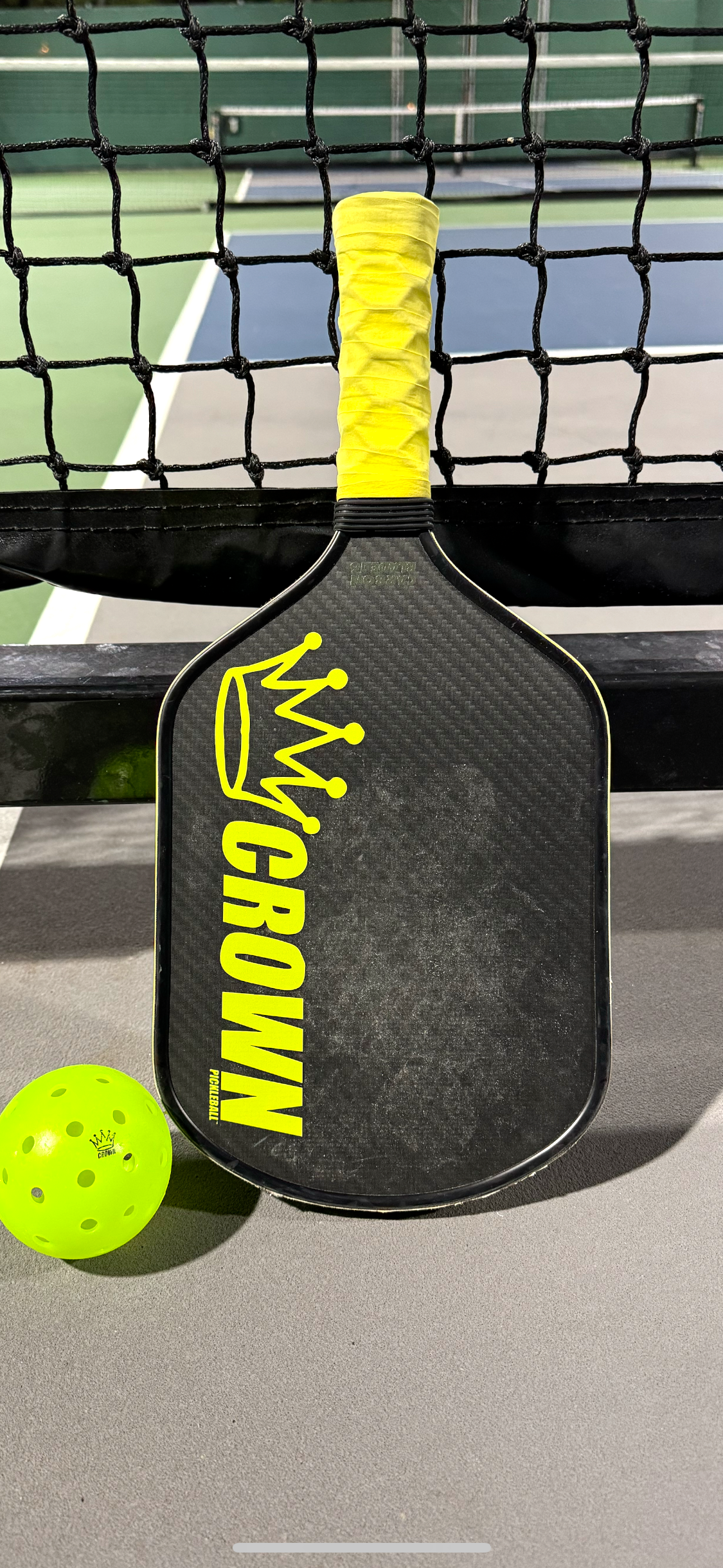 A CROWN PICKLEBALL - Carbon Blade 16 (BLACK Paddle) by Crown Pickleball, featuring a black design, yellow grip, and bold yellow “CROWN” text, leans against a net beside a bright yellow pickleball on a gray court.