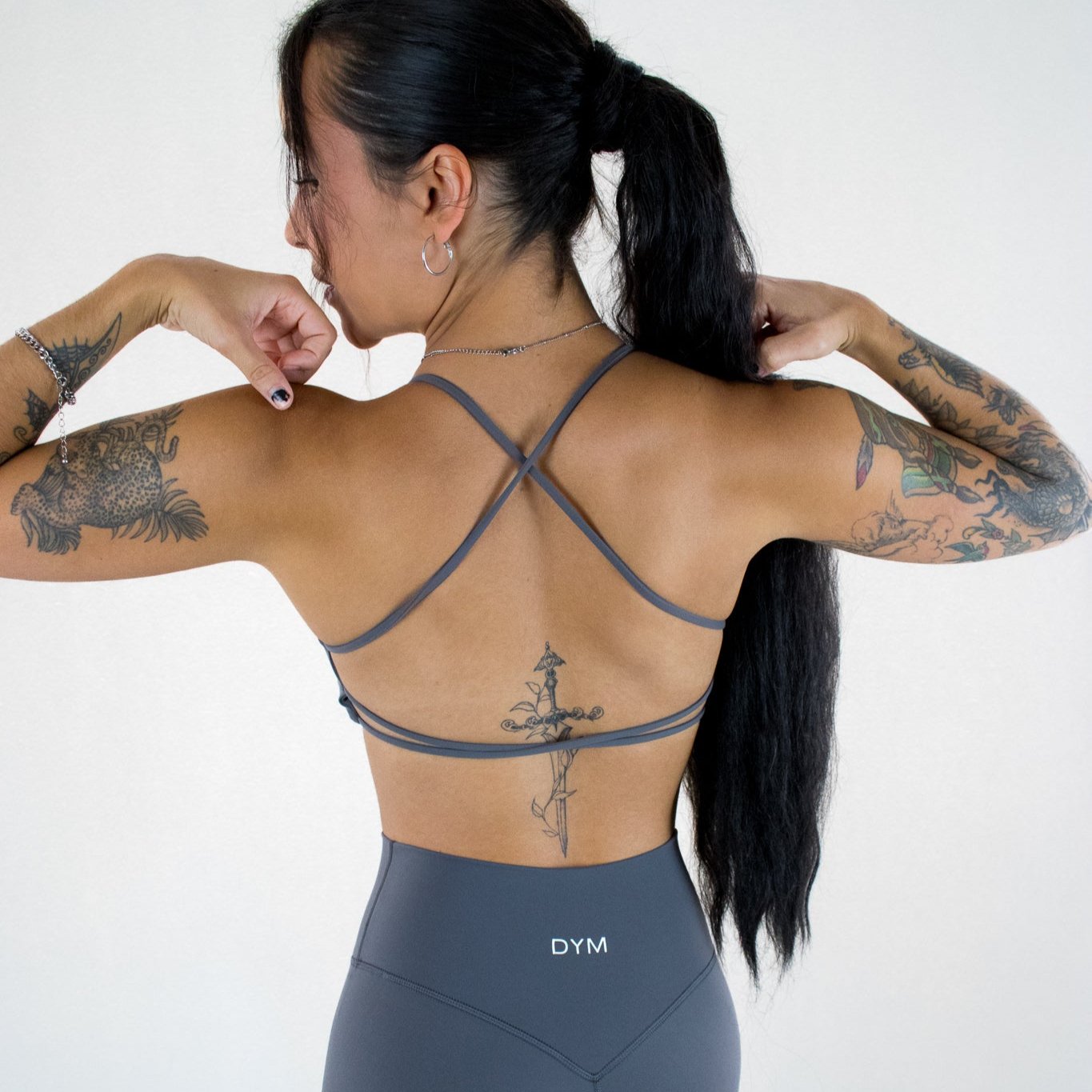 A woman with long black hair in a ponytail faces away, showing her back and tattooed arms. She wears the gray AIRE Ultra-Soft Open Back Bra by DYM Athletics with crisscross straps and matching leggings against a white background.
