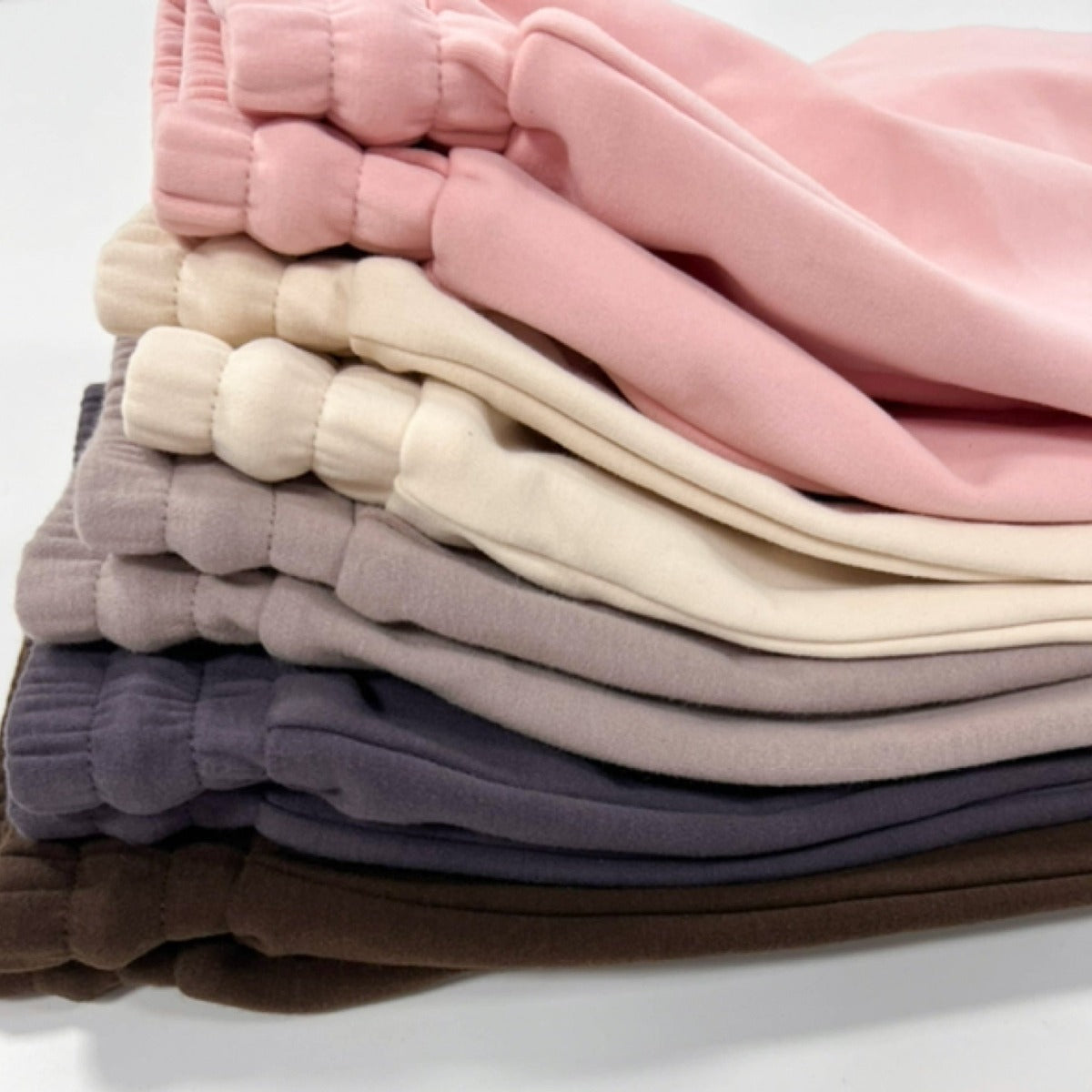 A neatly stacked pile of DYM Athletics Cozy Up Sweat Shorts in pink, cream, light taupe, gray, and brown with elastic waistbands arranged in a light-to-dark gradient on a white surface.