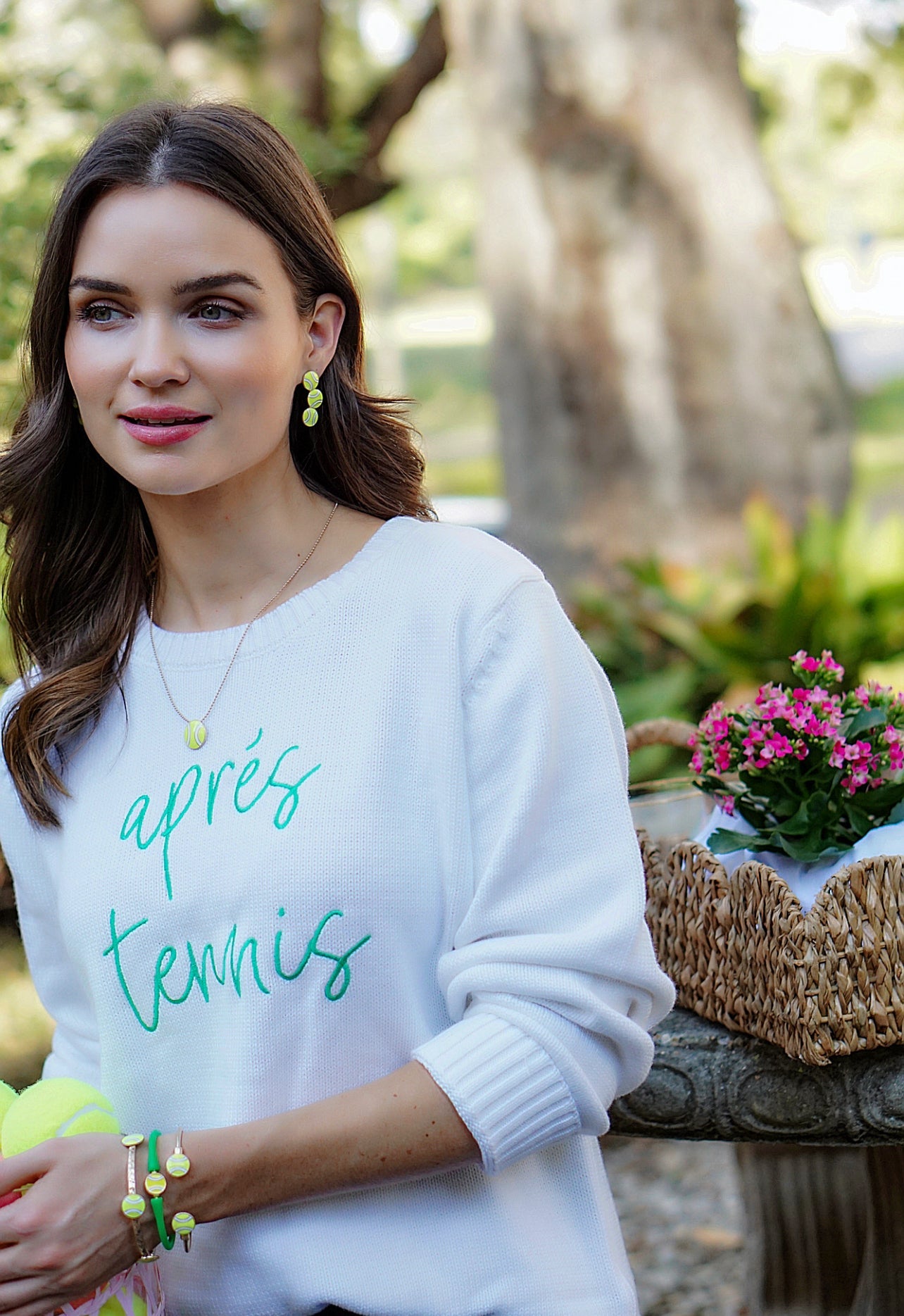 A woman with long brown hair wears the Runway Athletics Apres Tennis Sweater in white with green script, holding tennis balls and accessorized with yellow jewelry. A basket of pink flowers rests behind her among sunlit park trees—a perfect tennis gift.