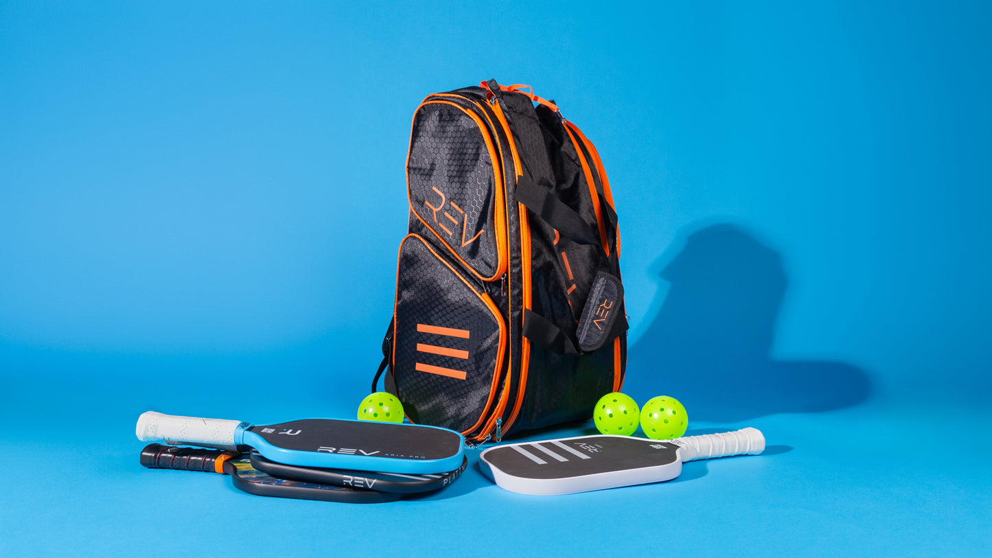 The REV Pickleball PRO Player Pickleball Bag, in black and orange, stands upright on a blue background, holding three paddles with white or black handles and unique designs, plus three neon yellow pickleballs—each fitting easily in insulated compartments.
