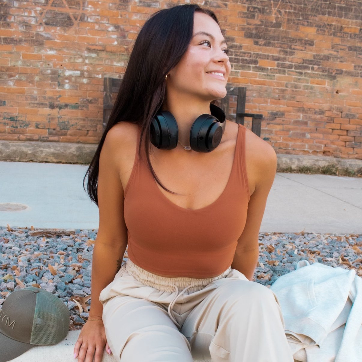 A smiling woman with long dark hair sits outside on rocks near a brick wall, wearing beige pants, black headphones around her neck, and a brown sleeveless top that reveals the buttery soft Flex V-Neck Bra with Scoop Back by DYM Athletics underneath.