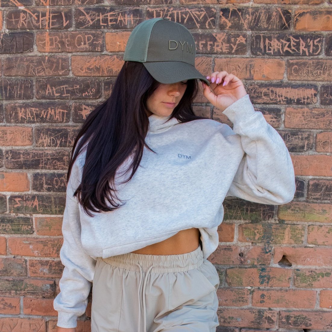 Standing against a graffiti-covered brick wall, a person with long dark hair looks down, holding the brim of their DYM Athletics | Trucker Hat. They wear a loose light gray cropped sweatshirt and beige pants.