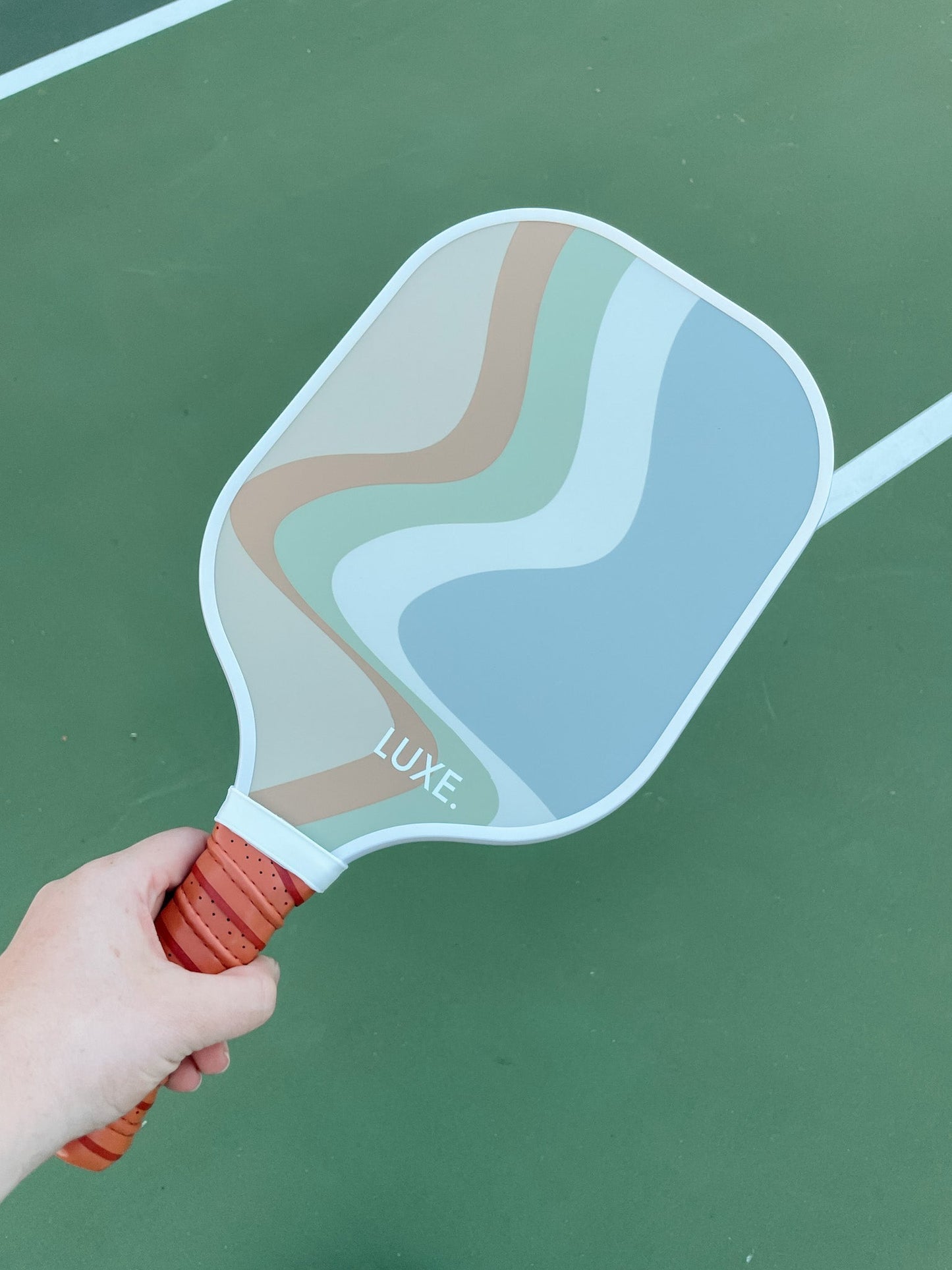 A hand holds The Luka paddle by LUXE Pickleball, featuring a pastel wavy design in blue, green, beige, and tan with a reddish-brown grip—perfect for players aiming to elevate their game on the outdoor court.
