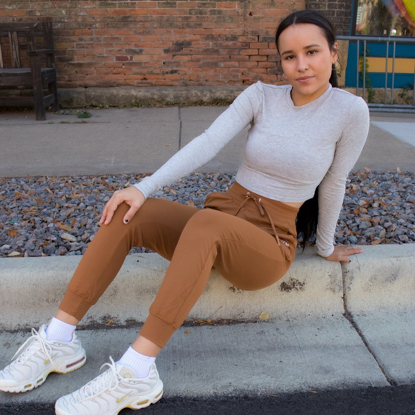 A young woman with long dark hair wears DYM Athletics Performance Pro Lightweight Active Jogger, a light gray long-sleeve shirt, and white sneakers as she sits on a curb. A gravel area, brick wall, and sidewalk provide the backdrop.