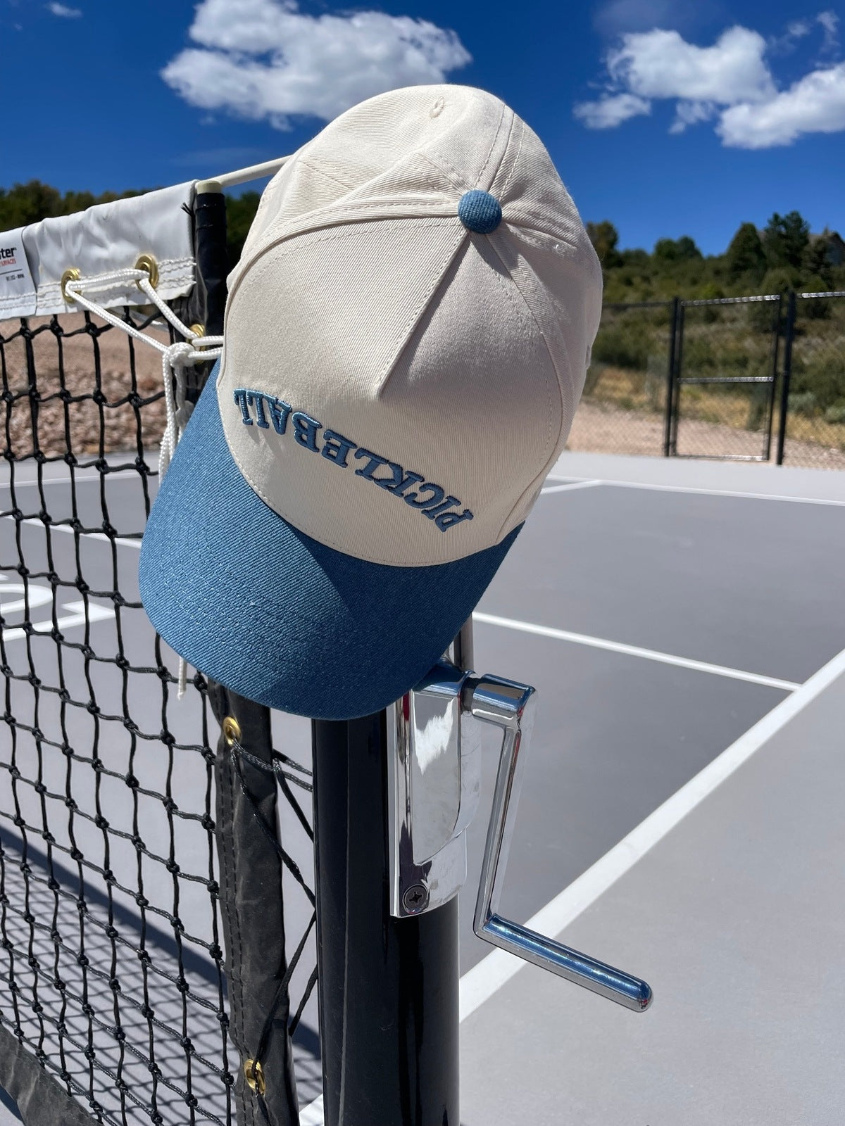 A beige and blue LUXE Pickleball Hat hangs on a silver net crank above the gray court, white lines, and black net. This Pickleball Hat offers sun protection with trees and a blue sky in the background.