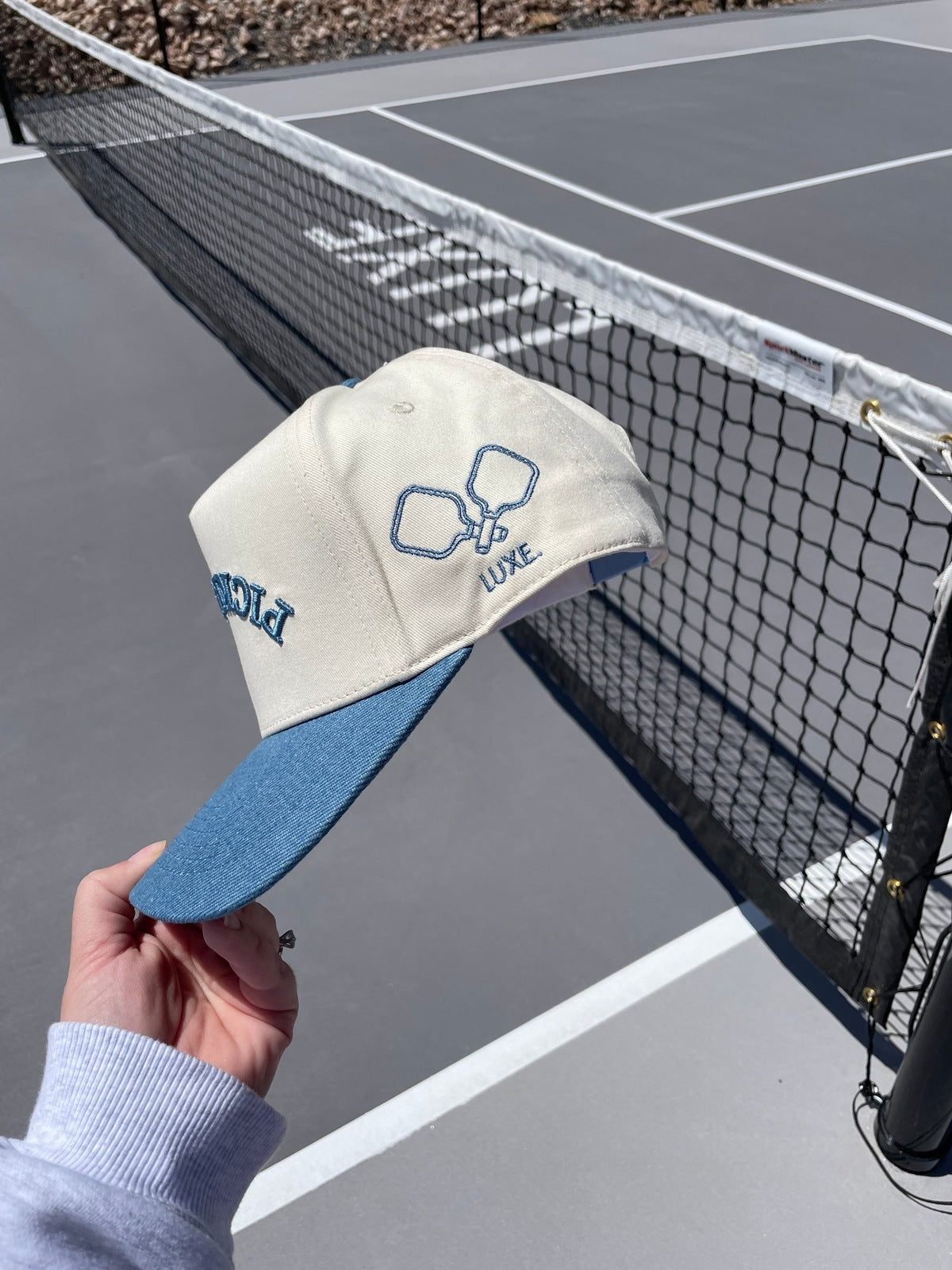 A person holds a beige and blue LUXE Pickleball Pickleball Hat with embroidered rackets and “LUXE” on the side by a black tennis net on a sunny court. The hat provides sun protection, with the net and court visible in the background.