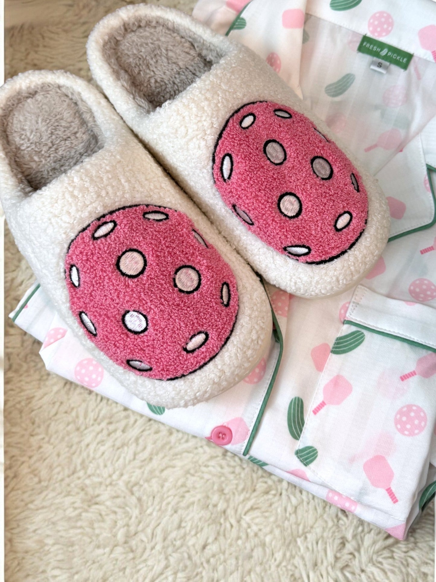 The Fresh Pickle Pickleball Slippers, featuring pink and white strawberry designs, rest on pastel pajamas with pink and green strawberries and green piping, all placed atop a cream-colored textured rug.