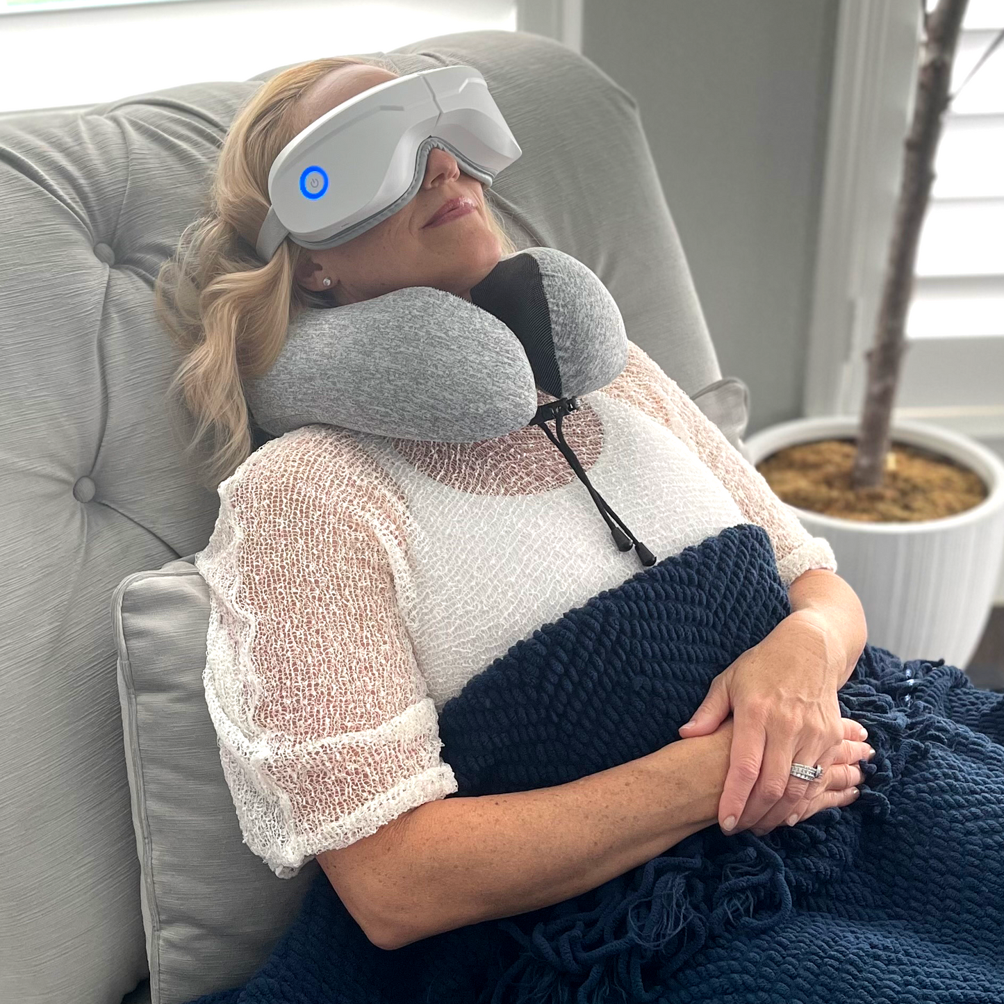 A woman reclines on a cushioned chair, enjoying the iReliev Relaxable™ Eye Massager with Heat featuring blue lights and compression therapy. She is wrapped in a dark blue knit blanket, with a potted plant in the background.