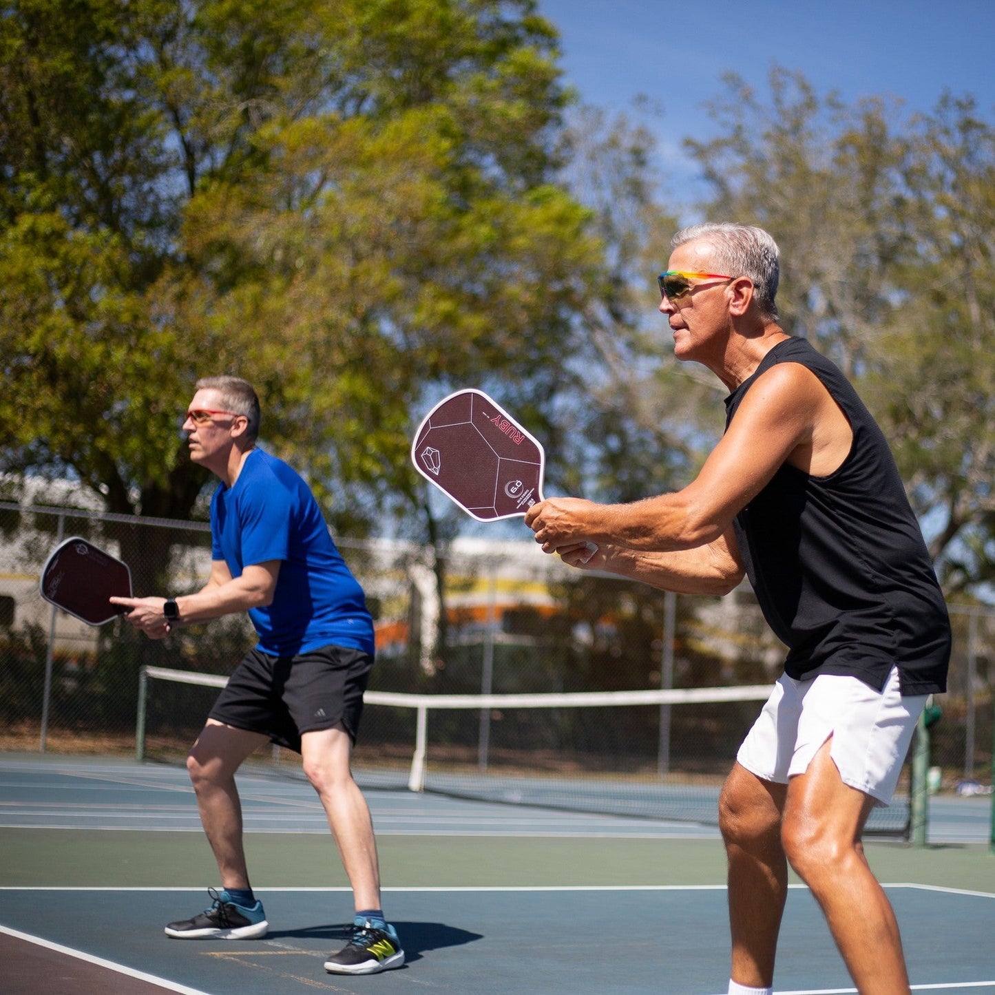 Two older men stand on an outdoor pickleball court, each holding a paddle and wearing Dink Eyewear Captiva Pink Pearl Pickleball Sunglasses, focusing on the game under a sunny sky with trees in the background.