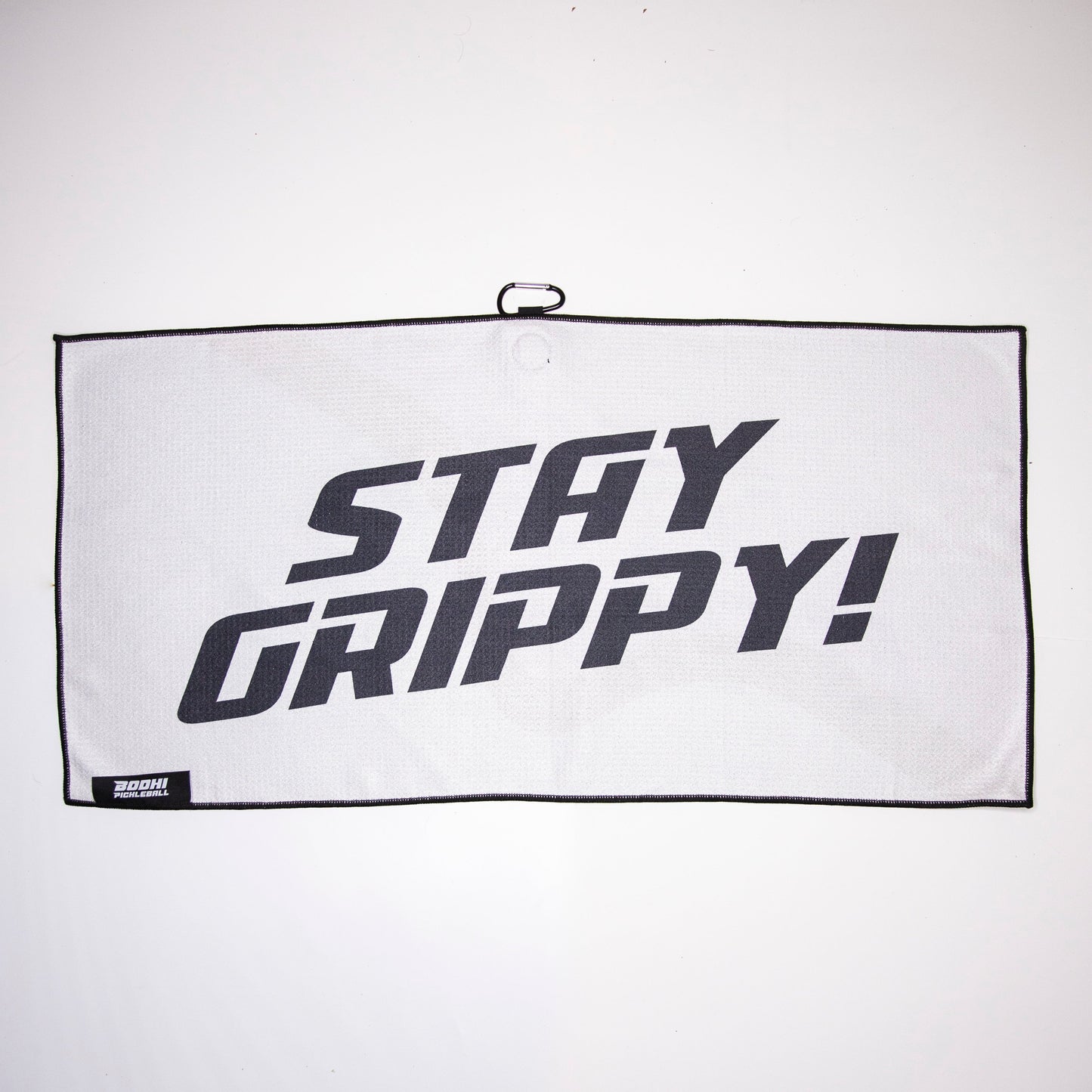 The white, rectangular Bodhi Performance Pickleball Towel with black trim features bold “STAY GRIPPY!” text in the center, a small label at the bottom left, and a hanging loop for easy storage among your gear.