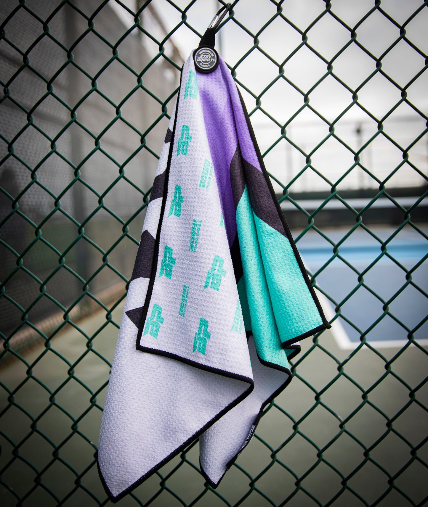 The Bodhi Performance Performance Pickleball Towel, with teal, purple, and black geometric patterns, hangs by a black carabiner on a chain-link fence near a tennis court under an overcast sky.