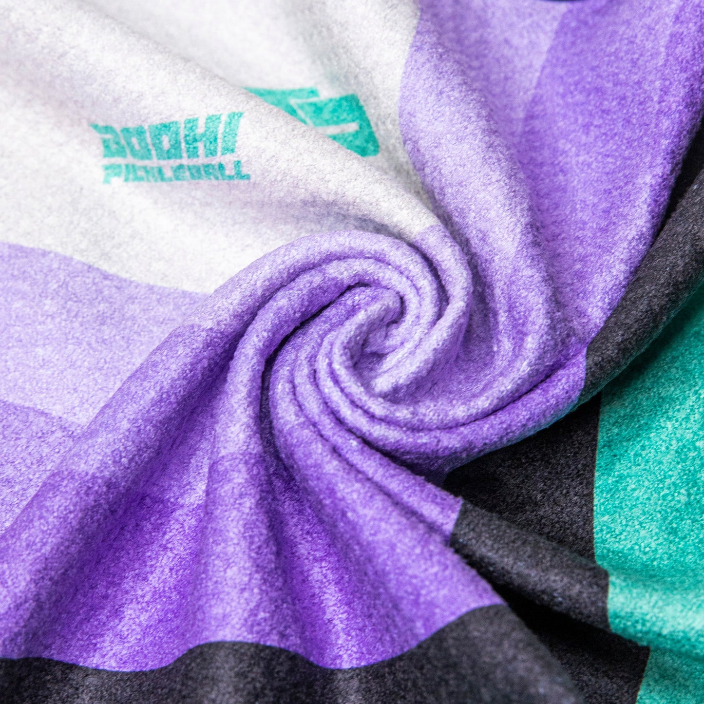 A close-up of Bodhi Performance’s Performance Pickleball Towel, swirled to show soft microfiber fabric with bold black, dark green, purple, light purple, and white stripes plus a teal 30HHI PICKLEBALL logo—a must-have pickleball accessory.